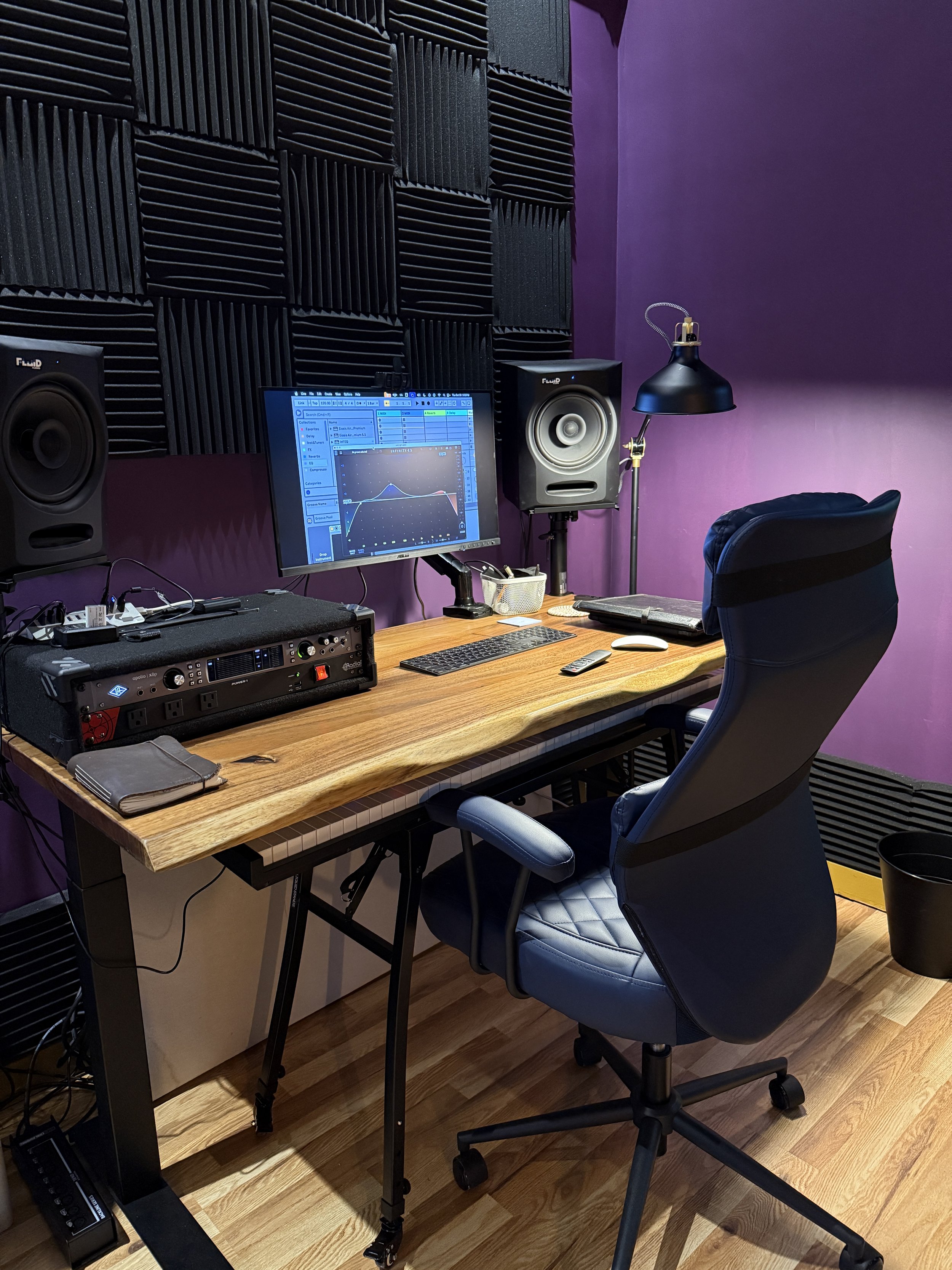 A recording or music production studio workspace with a wooden desk, a computer monitor displaying an audio editing software, studio monitor speakers, acoustic foam panels on the purple walls, a black ergonomic office chair, a desk lamp, and various 