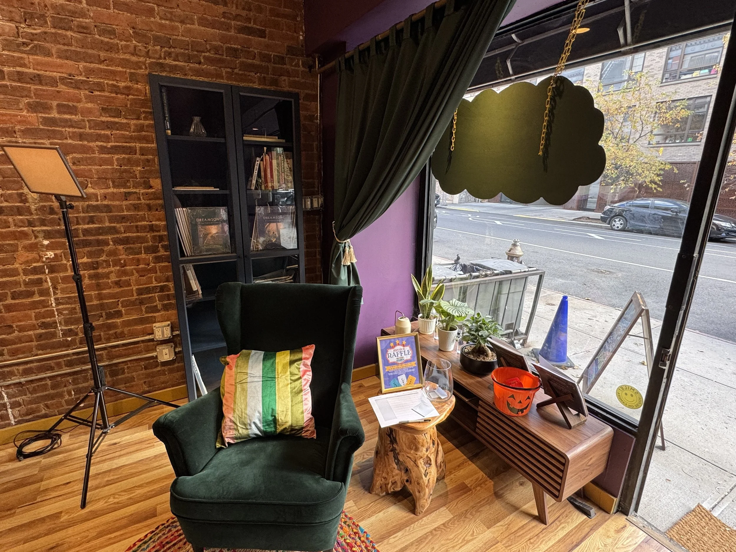 Cozy cafe interior with a green armchair, a colorful pillow, a wooden side table with a glass of wine, and a plant in a pot near a large open window covered partly by green curtains. Outside visible are cars, a street, and trees.