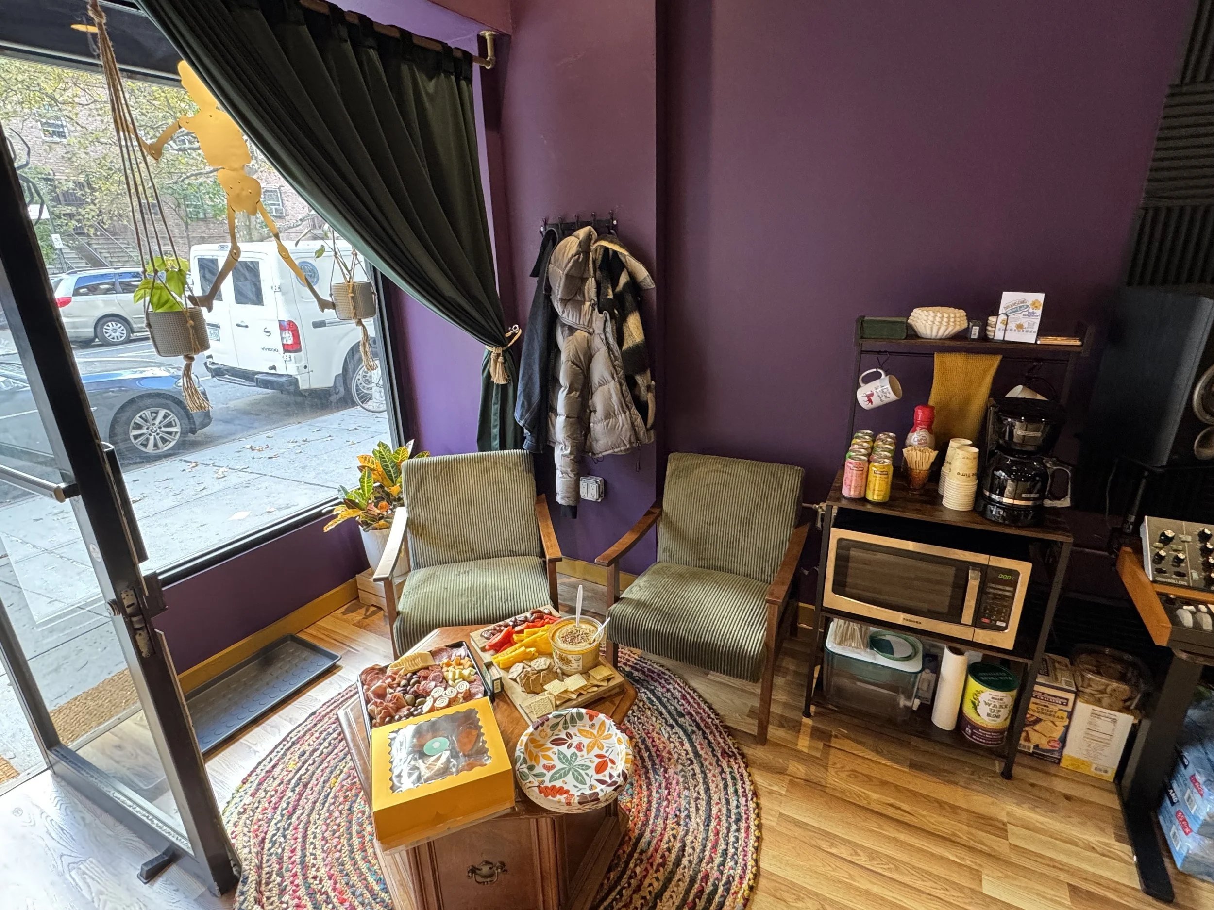 A cozy corner of a café or home with a purple wall, green chairs, a small wooden table with snacks, and a window with plants and hanging decorations outside.