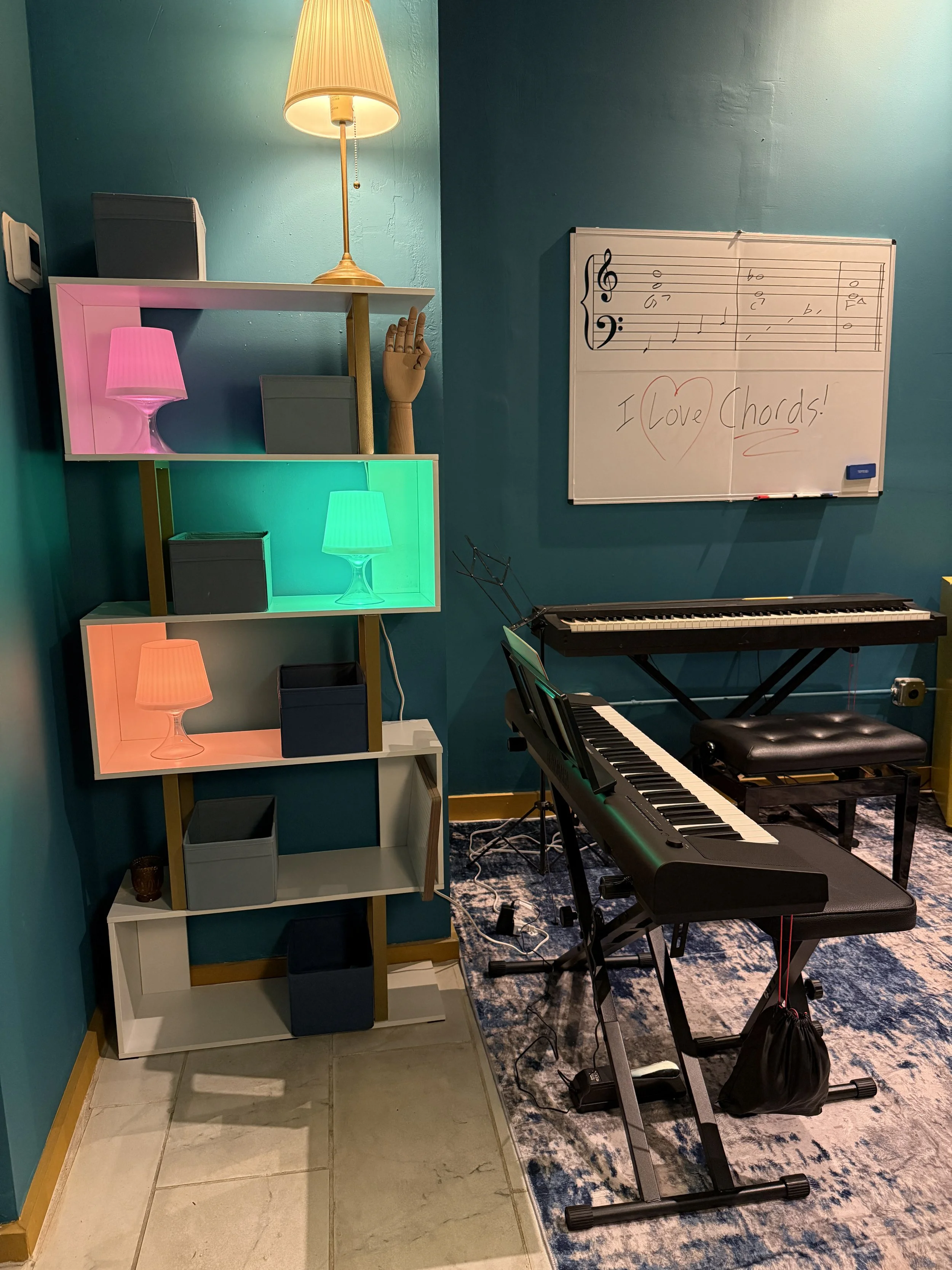 A music practice room featuring a digital keyboard, an electronic keyboard on a stand, a whiteboard with musical notes and a message, a shelf with colorful lamps, a wooden mannequin hand, and a lamp with a yellow shade.