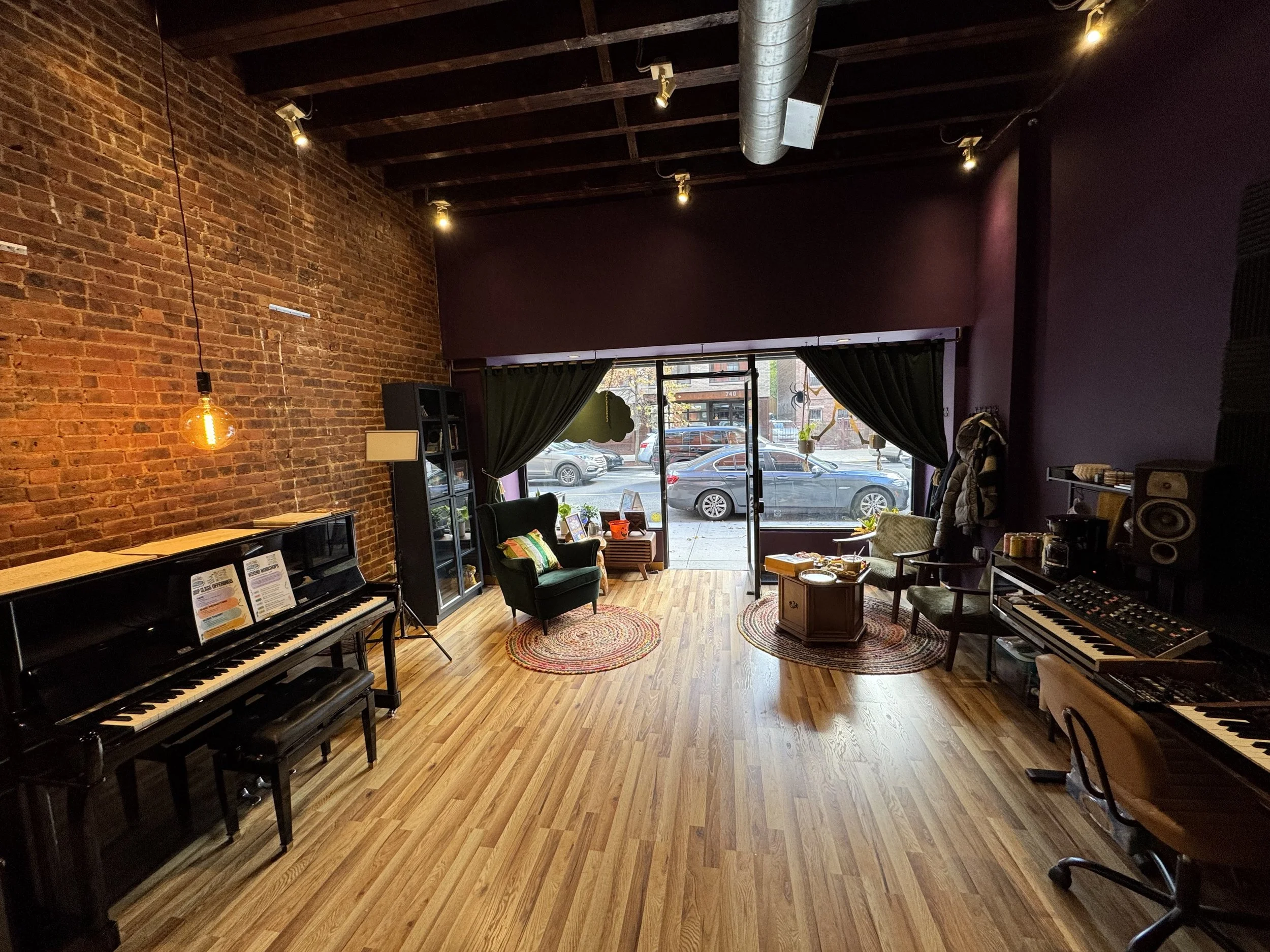 Interior view of a cozy music studio with brick walls, hardwood floors, and various musical instruments including a piano and keyboard. There is seating near large windows with black curtains, and sunlight outside illuminates the room.