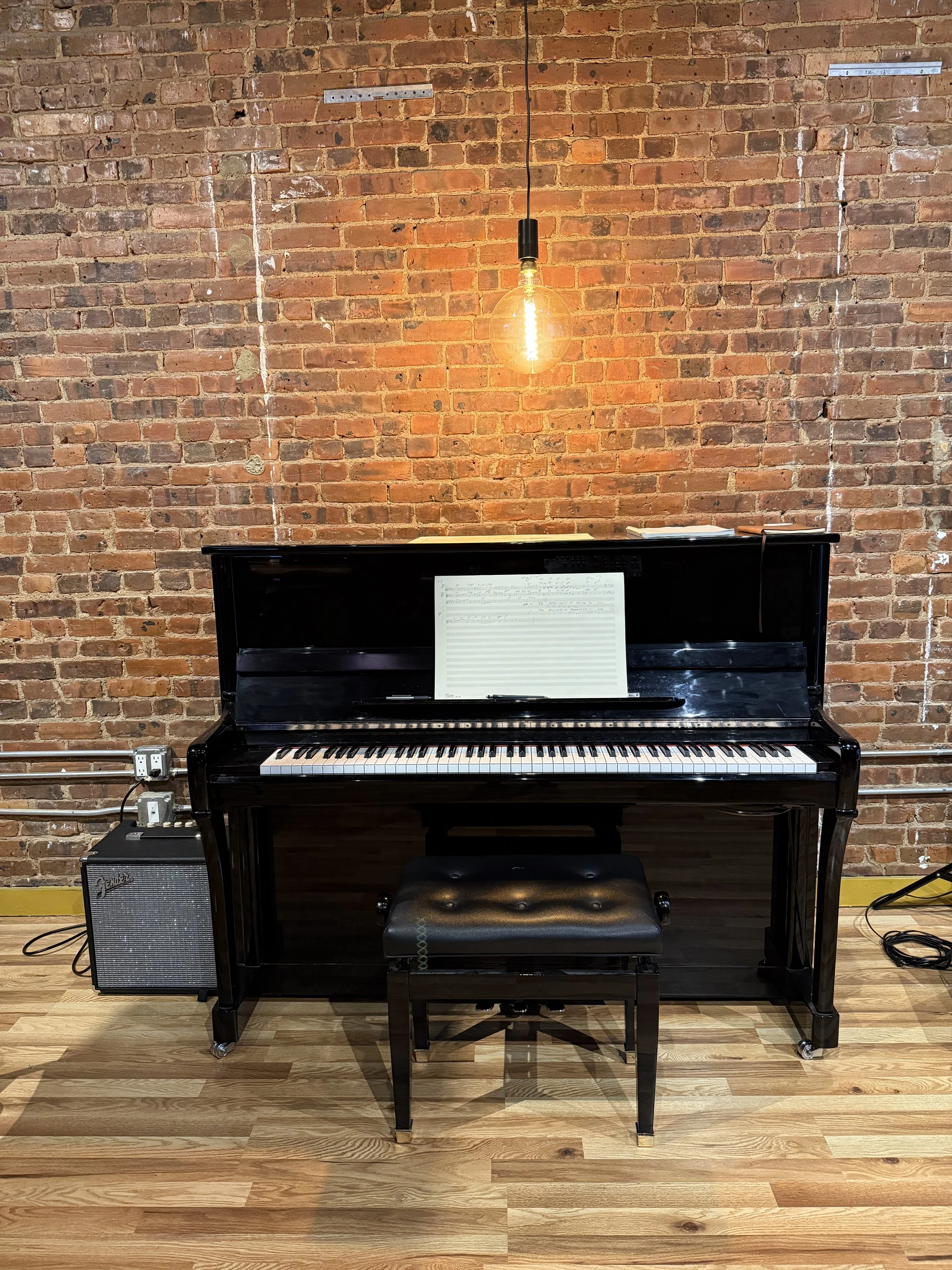 A black upright piano with an open sheet music stand, placed against an exposed brick wall. A small bench with a black cushioned top is in front of the piano. To the left of the piano, there is a small amplifier connected to a power strip, and a hang