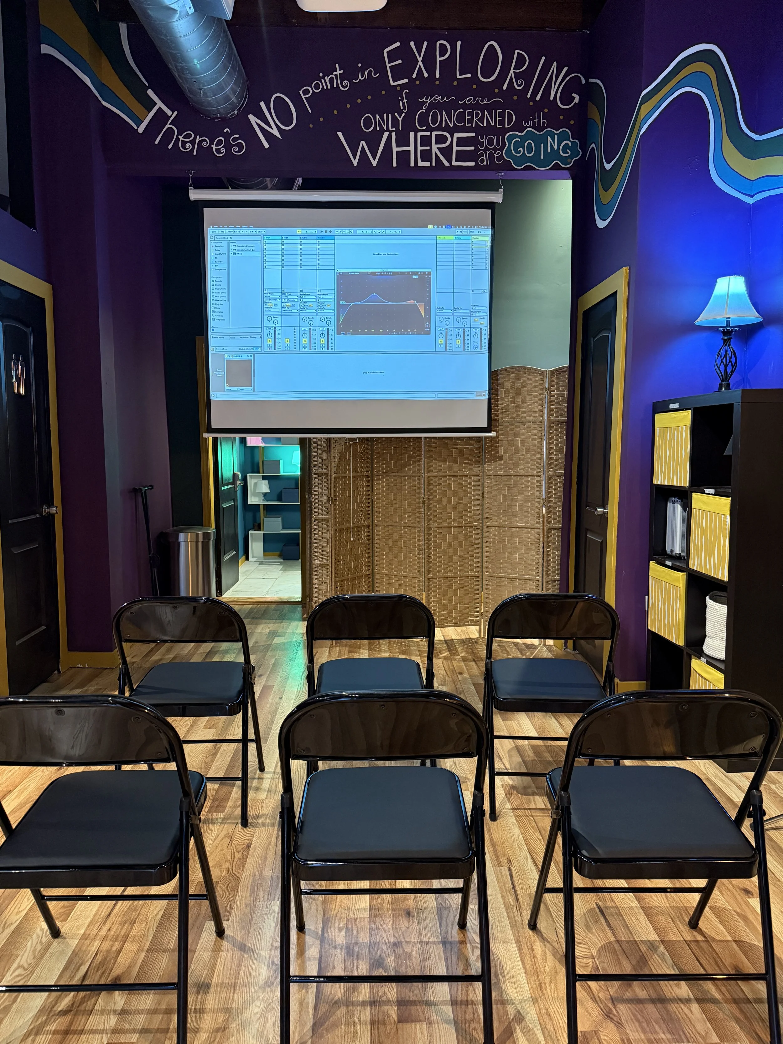 A small indoor room set up for a presentation or workshop with six black chairs facing a projector screen displaying music production software. The room has purple walls decorated with a motivational quote, a black bookshelf with yellow and white sto