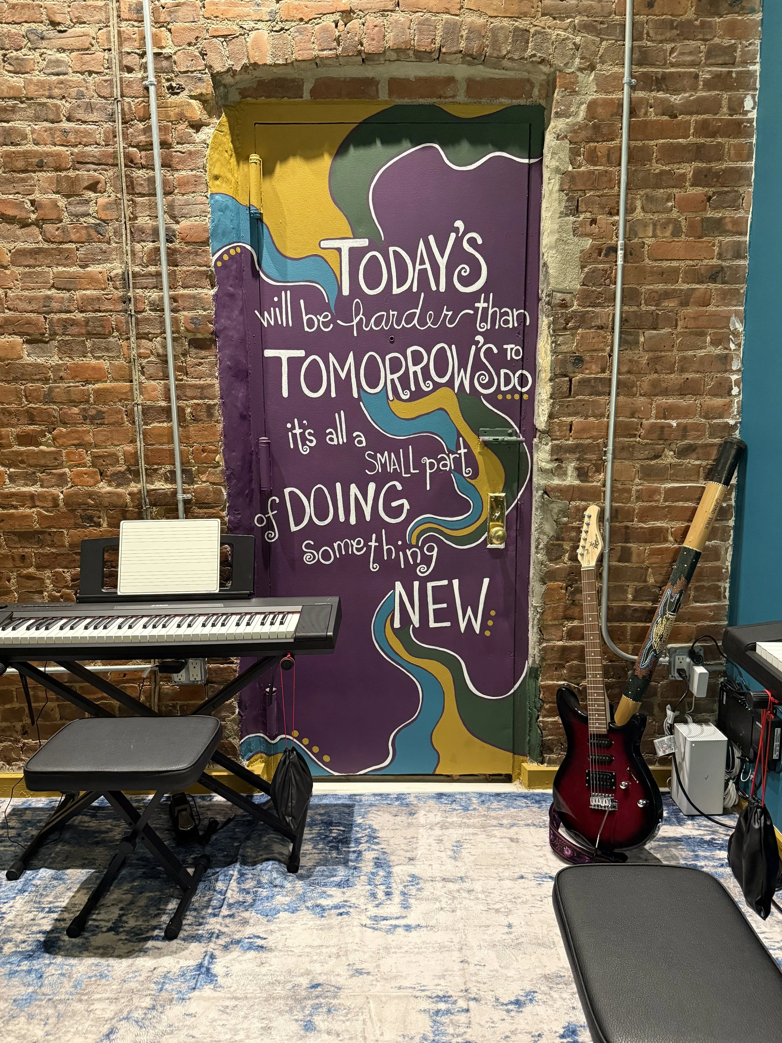 Colorful mural on brick wall with a motivational quote, musical keyboard on stand, chair, electric guitar, and a flute leaning against the wall.