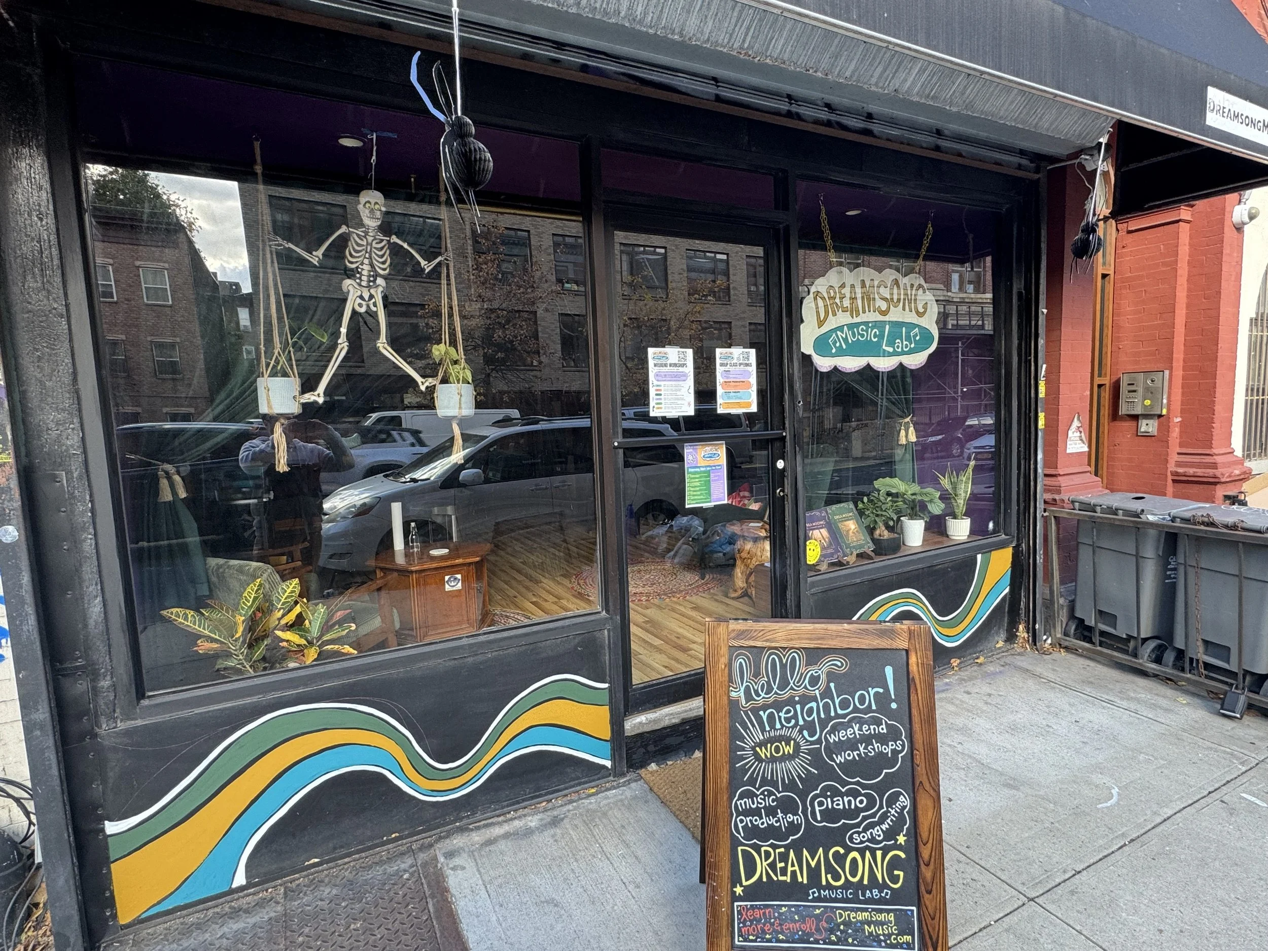The storefront of Dreamsong Music Lab with colorful wavy decoration at the bottom of the windows, a skeleton decoration hanging inside, and a chalkboard sign outside advertising weekend workshops for music production, piano, and songwriting.