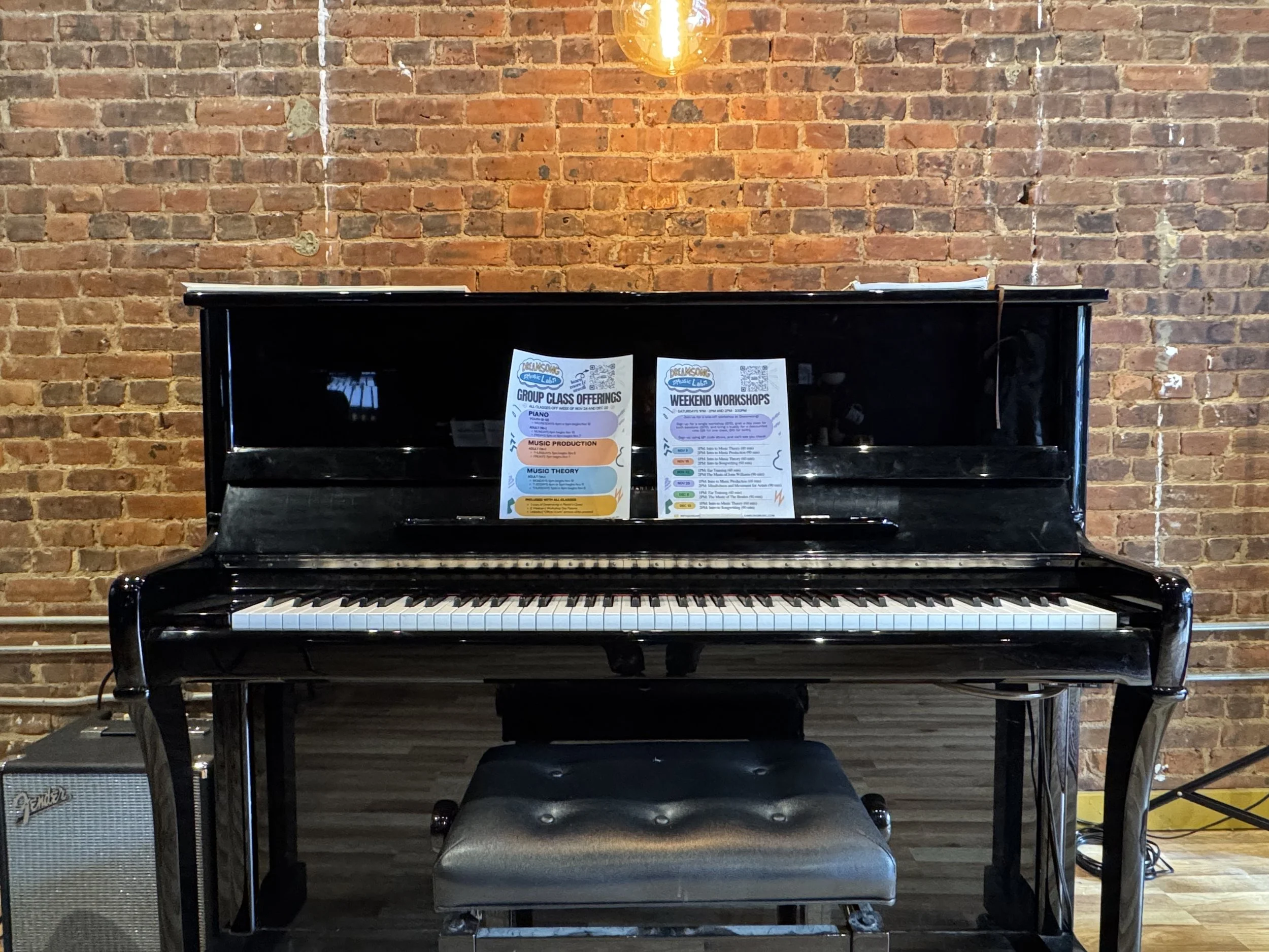 Black upright piano with two sheets of paper on music stand, against exposed brick wall, with a black cushioned bench in front, and a small Fender amplifier to the left.
