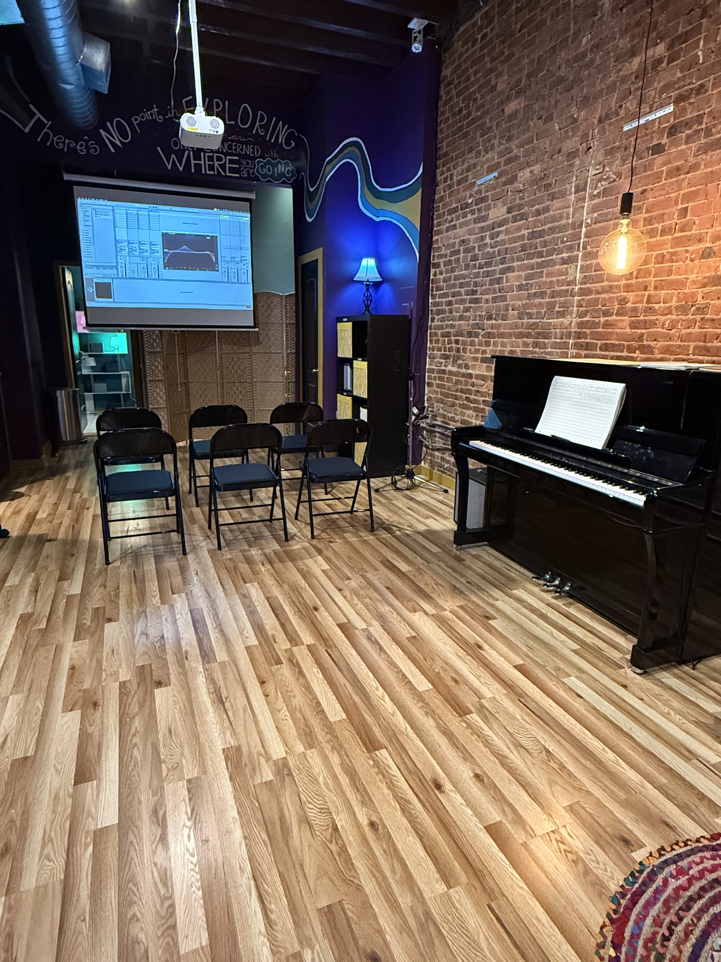 A small music room with a black upright piano, sheet music on top, six black chairs arranged in a semi-circle, a projector screen displaying music or audio software, a blue lamp, and colorful wall art on a dark purple wall with an inspiring quote abo
