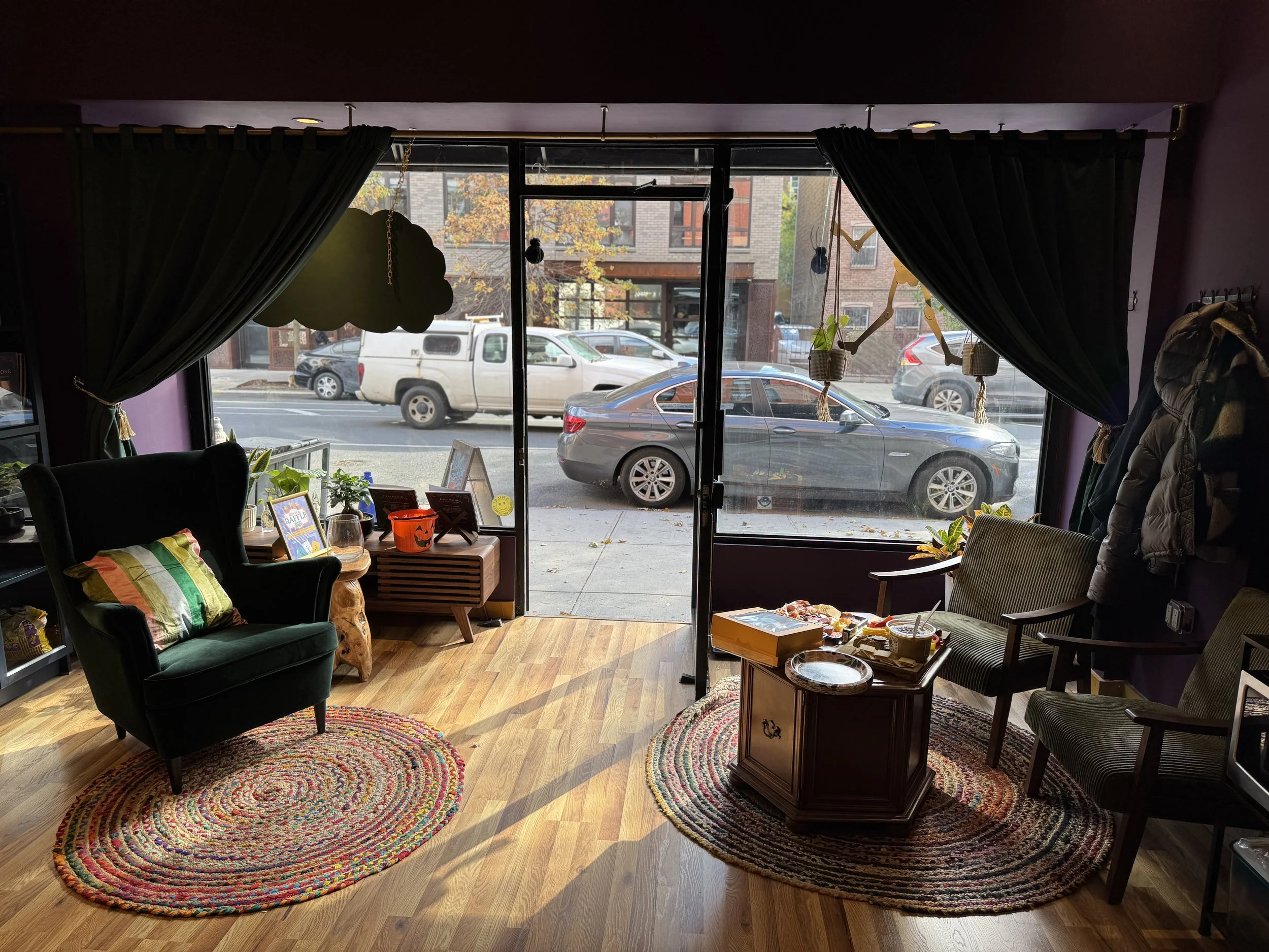 Cozy interior of a cafe or waiting area with a large window, black curtains, a colorful braided rug, a green velvet armchair with a striped pillow, two matching chairs, small tables, and decorations including plants and picture frames.