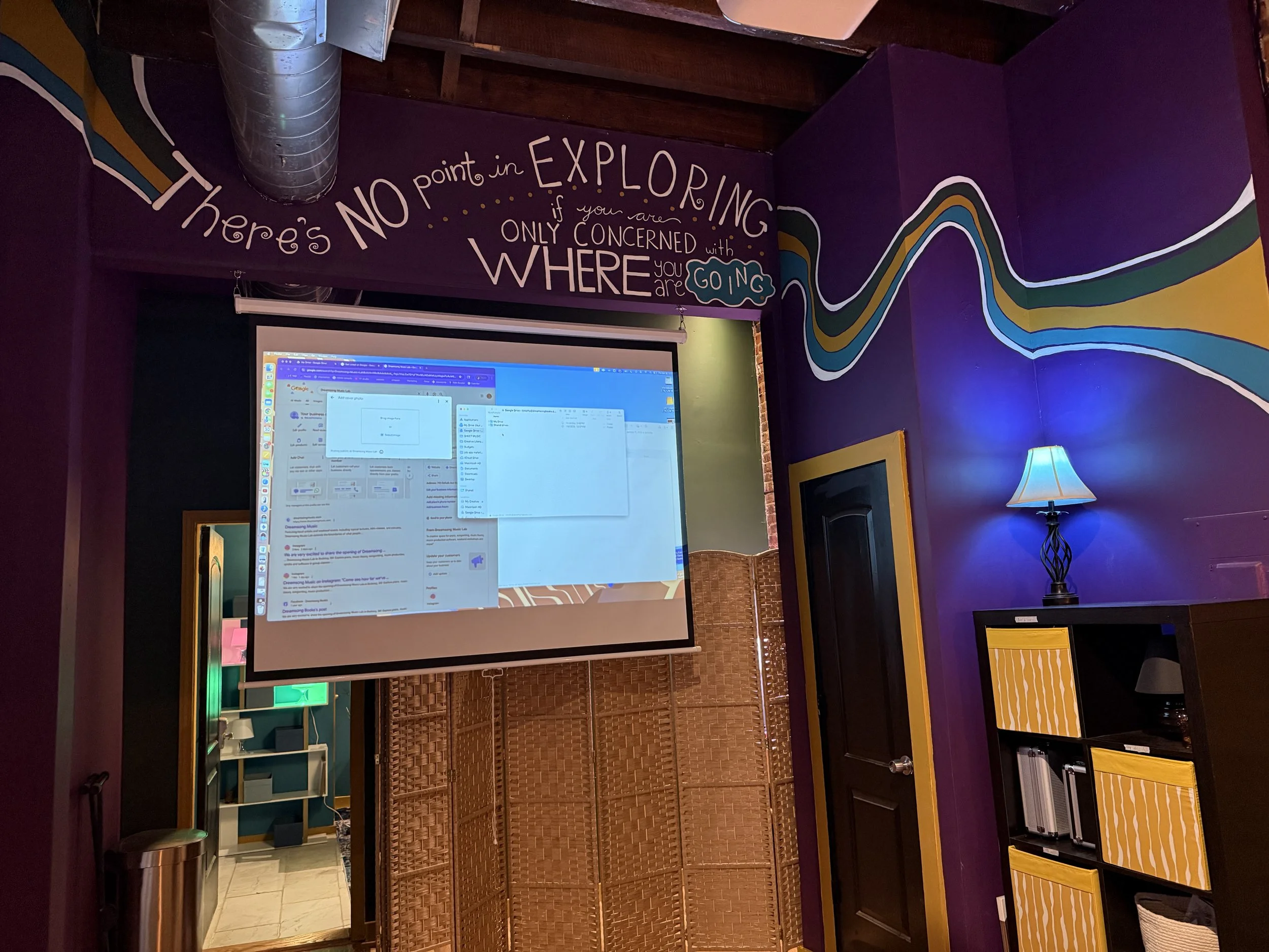 Colorful room with purple walls, a large projector screen showing computer windows, a small table with a lamp, and a bookshelf with yellow and black storage boxes. Artistic wall mural with text that says, 'There's NO point in EXPLORING if you are ONL