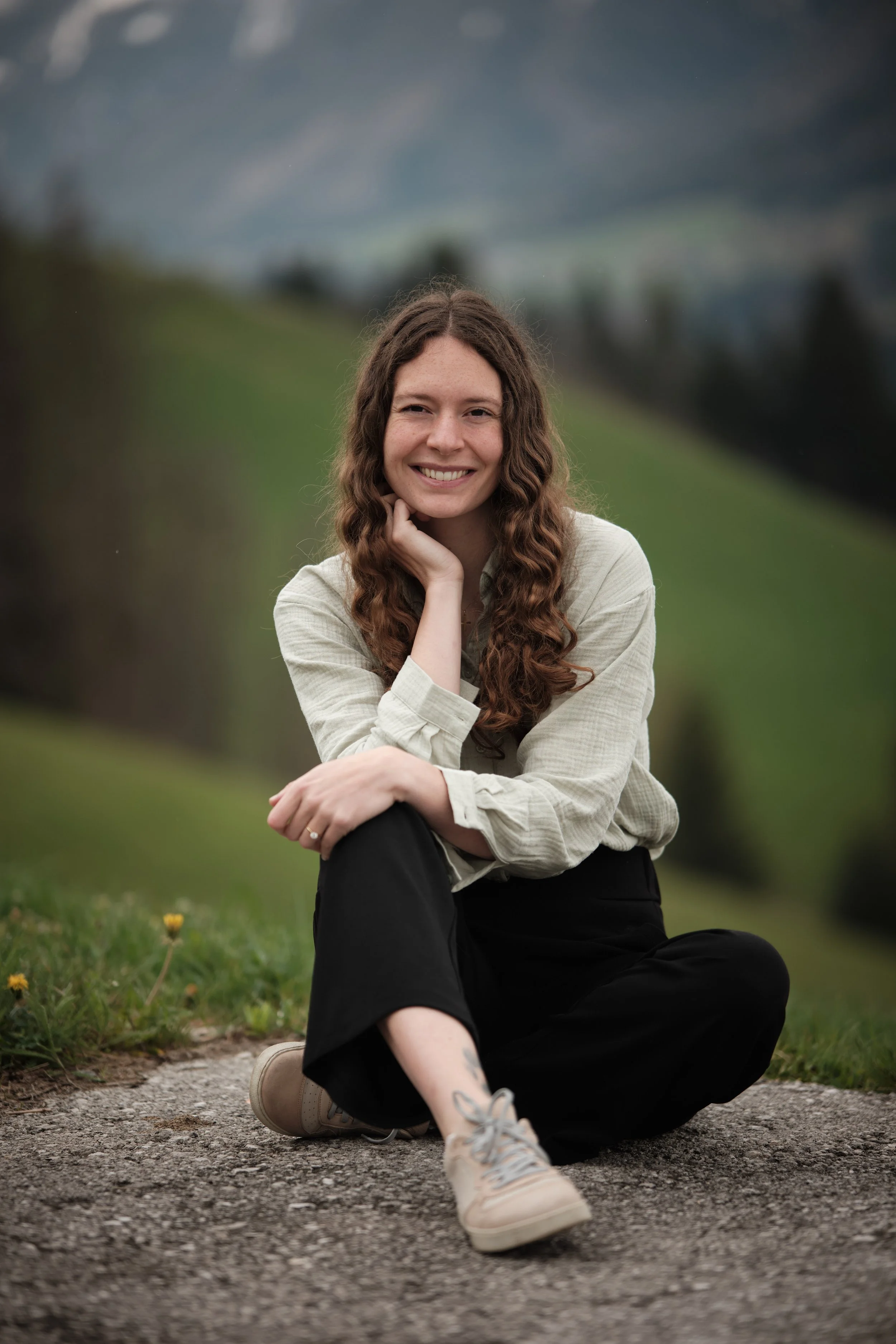 Eine lachende junge Frau mit lockigem braunem Haar sitzt auf einem Weg in einer grünen, hügeligen Landschaft mit fernen Bergen im Hintergrund.