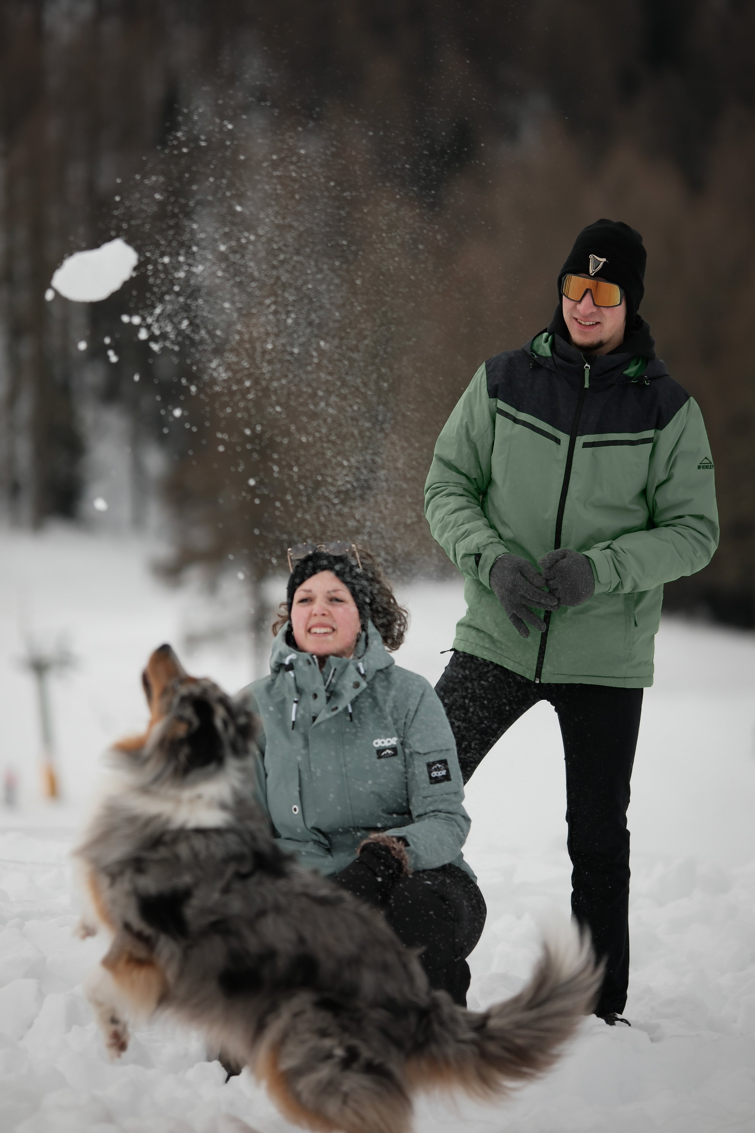 Zwei Menschen spielen im Schnee, ein Mann steht und ein Frau kniet, während ein Hund im Vordergrund herumläuft, mit verschneiter Landschaft im Hintergrund.