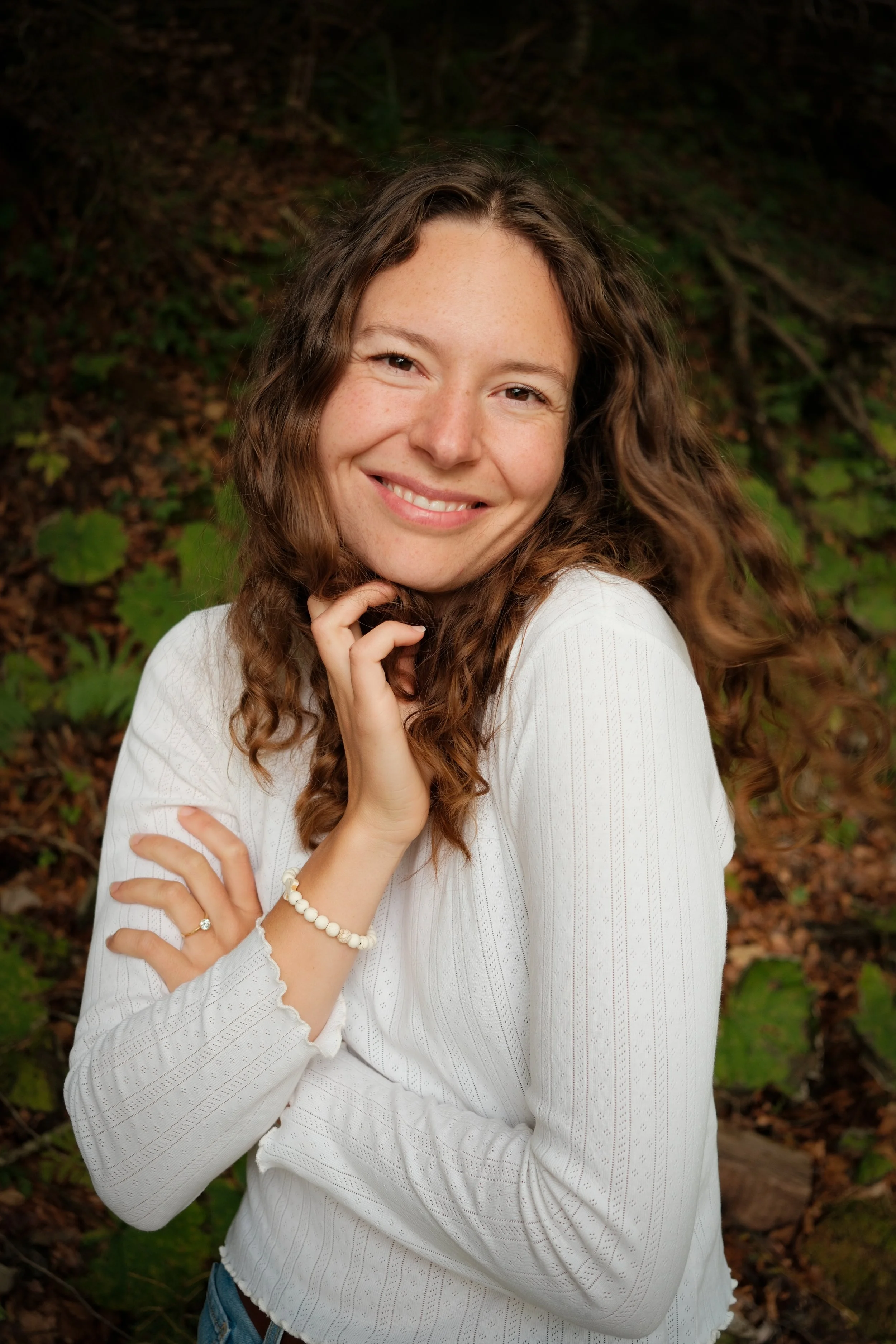 Lächelnde junge Frau mit lockigem Haar, weißem Oberteil und Perlenarmband, im Wald stehender Hintergrund.