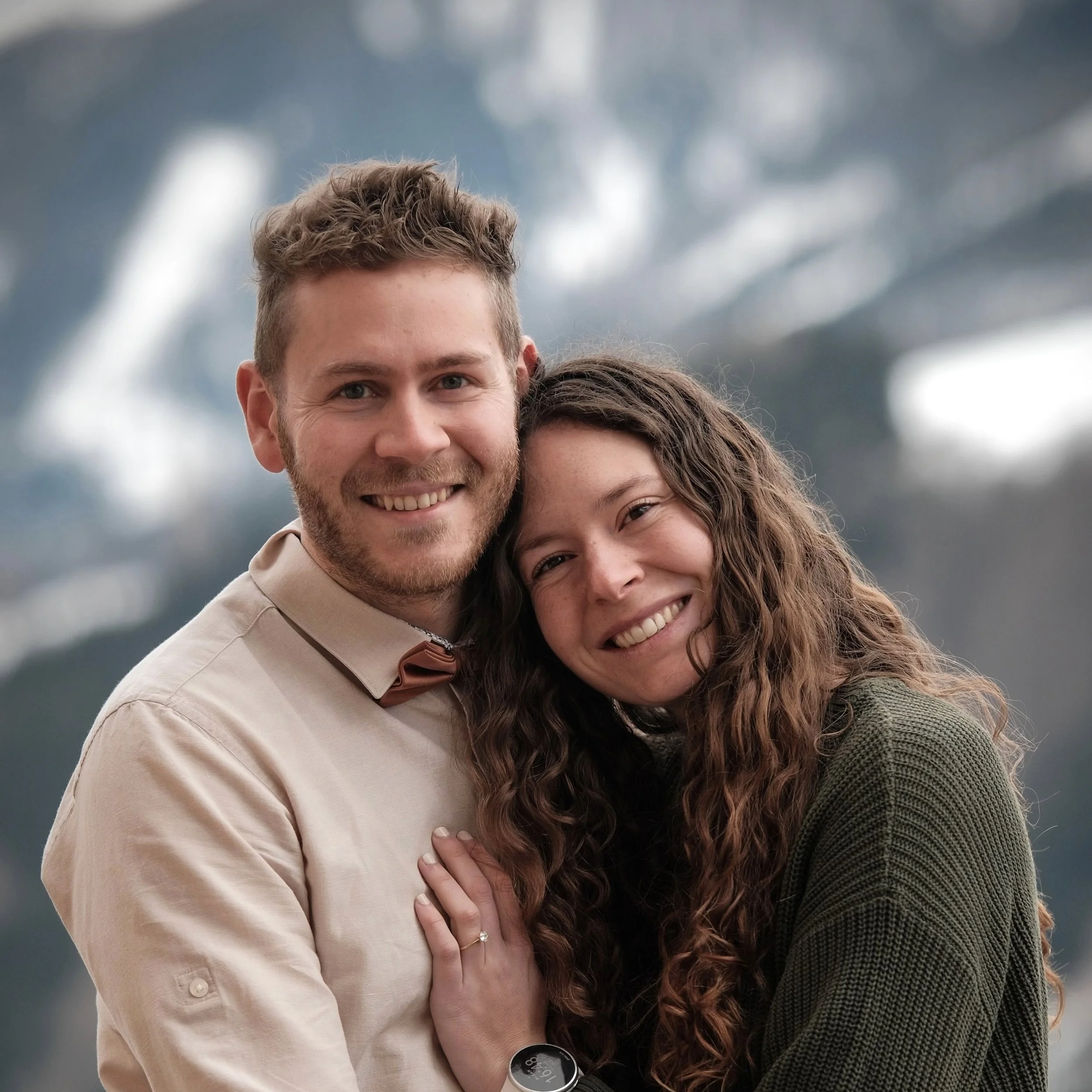 Ein glückliches Paar, das draußen vor einer Bergkulisse lächelt, umarmt sich und zeigt ihre Ringe.