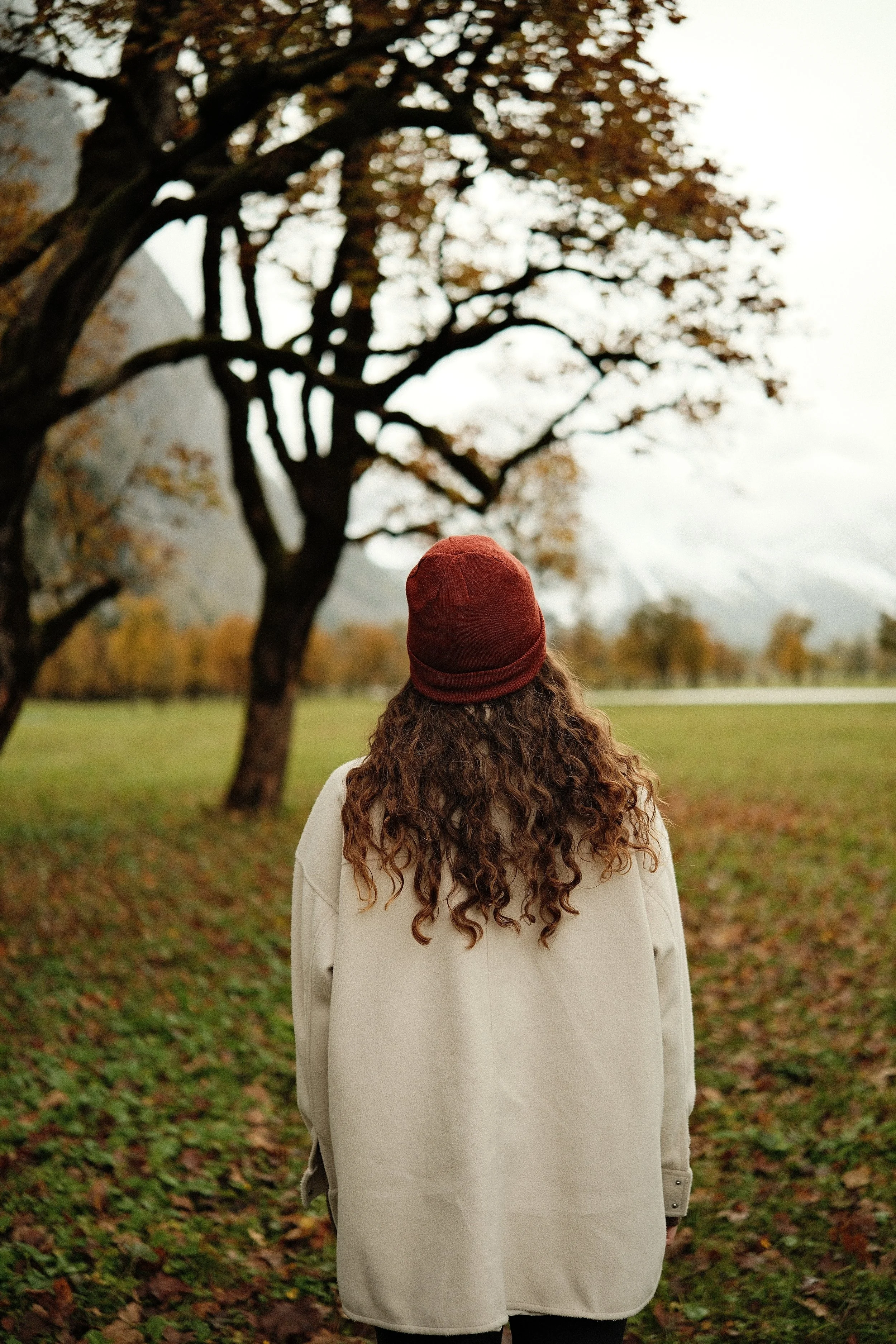 Eine Person mit lockigem Haar, die eine beige Jacke und eine braune Mütze trägt, steht in einem Park mit Bäumen im Herbst, Blick in die Ferne, Berg im Hintergrund.