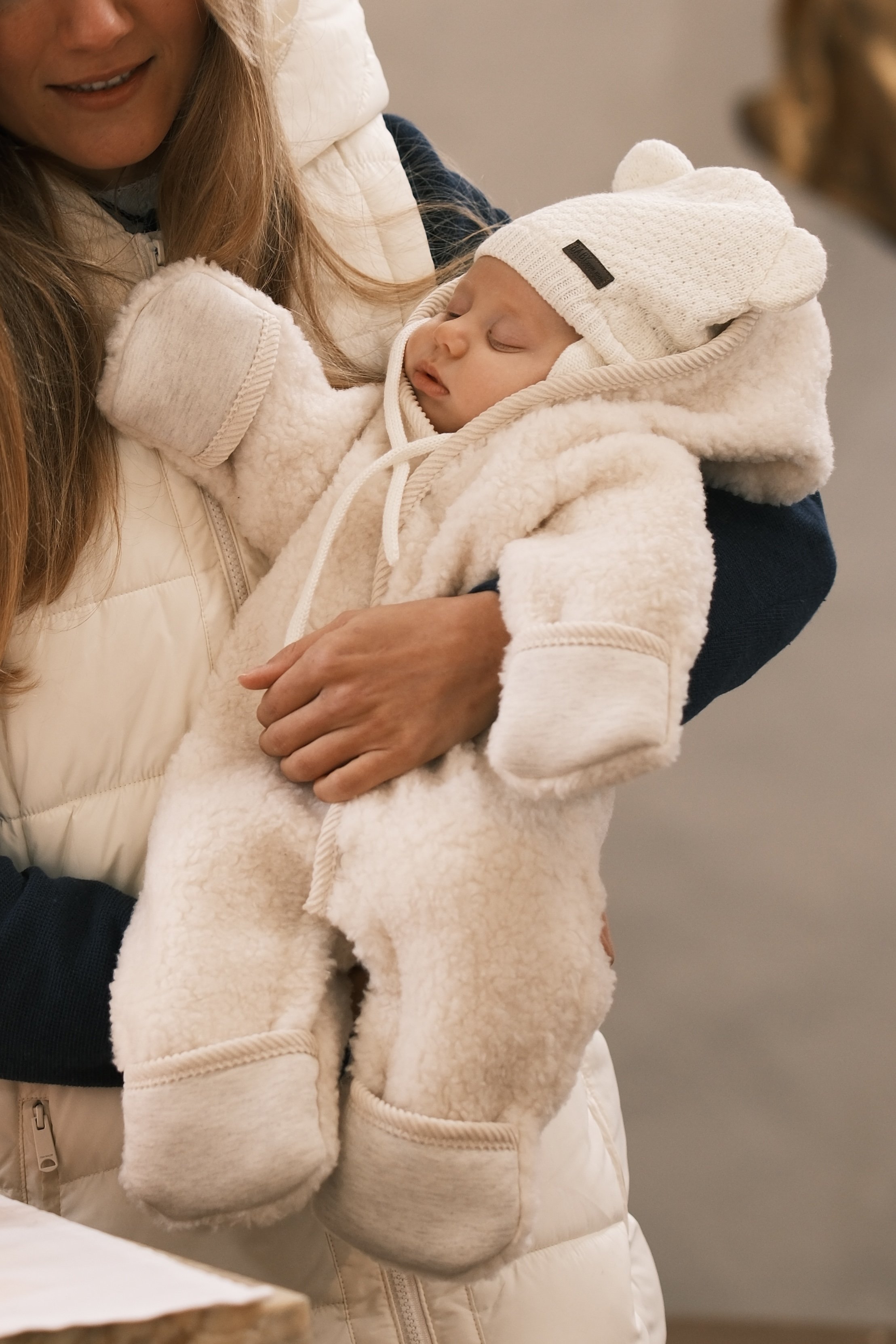 Ein Baby in einem weißen, kuscheligen Overall und einer Mütze, das in den Armen einer Frau schläft.