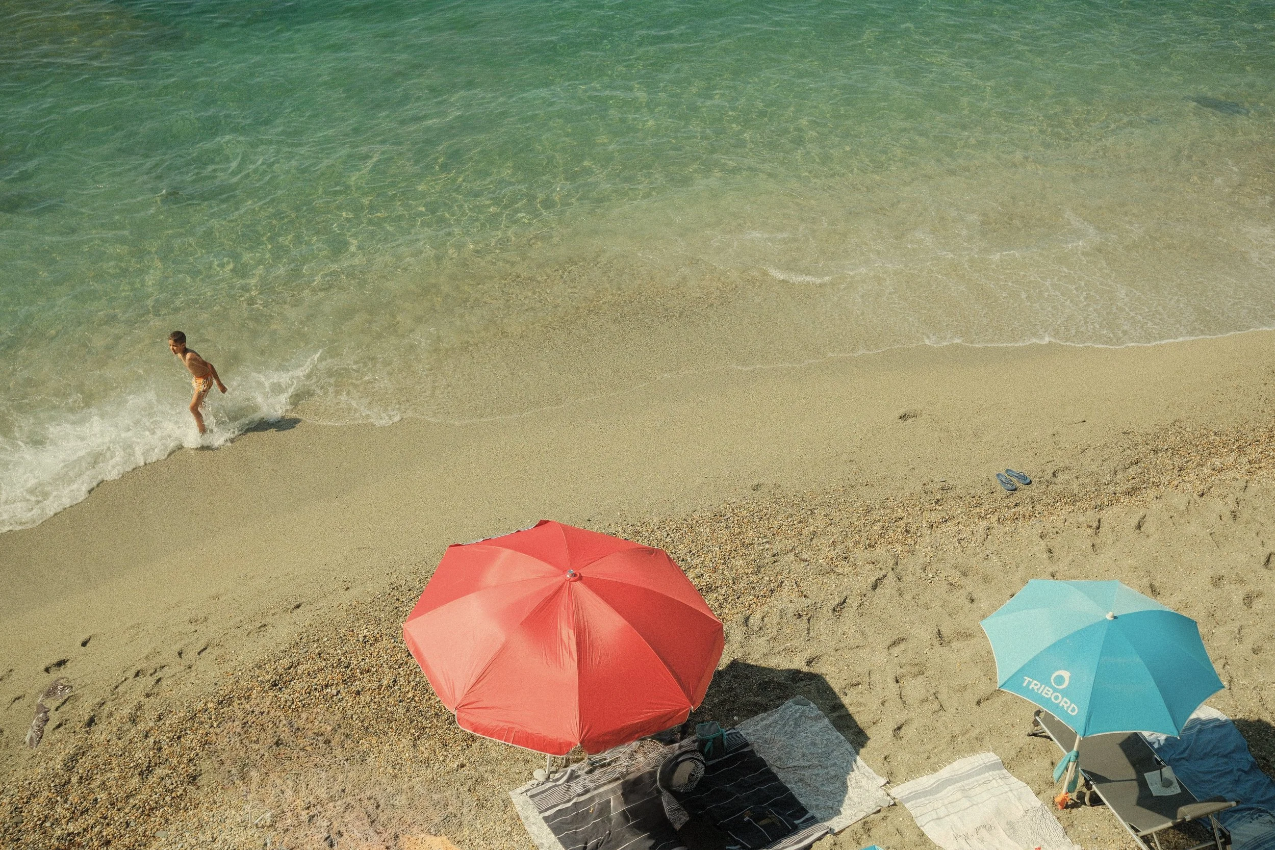 Strand mit einem Kind, das im Wasser spielt, zwei Sonnenschirme (rot und blau), Strandtücher, eine Sonnenbrille und Schuhe auf dem Sand.