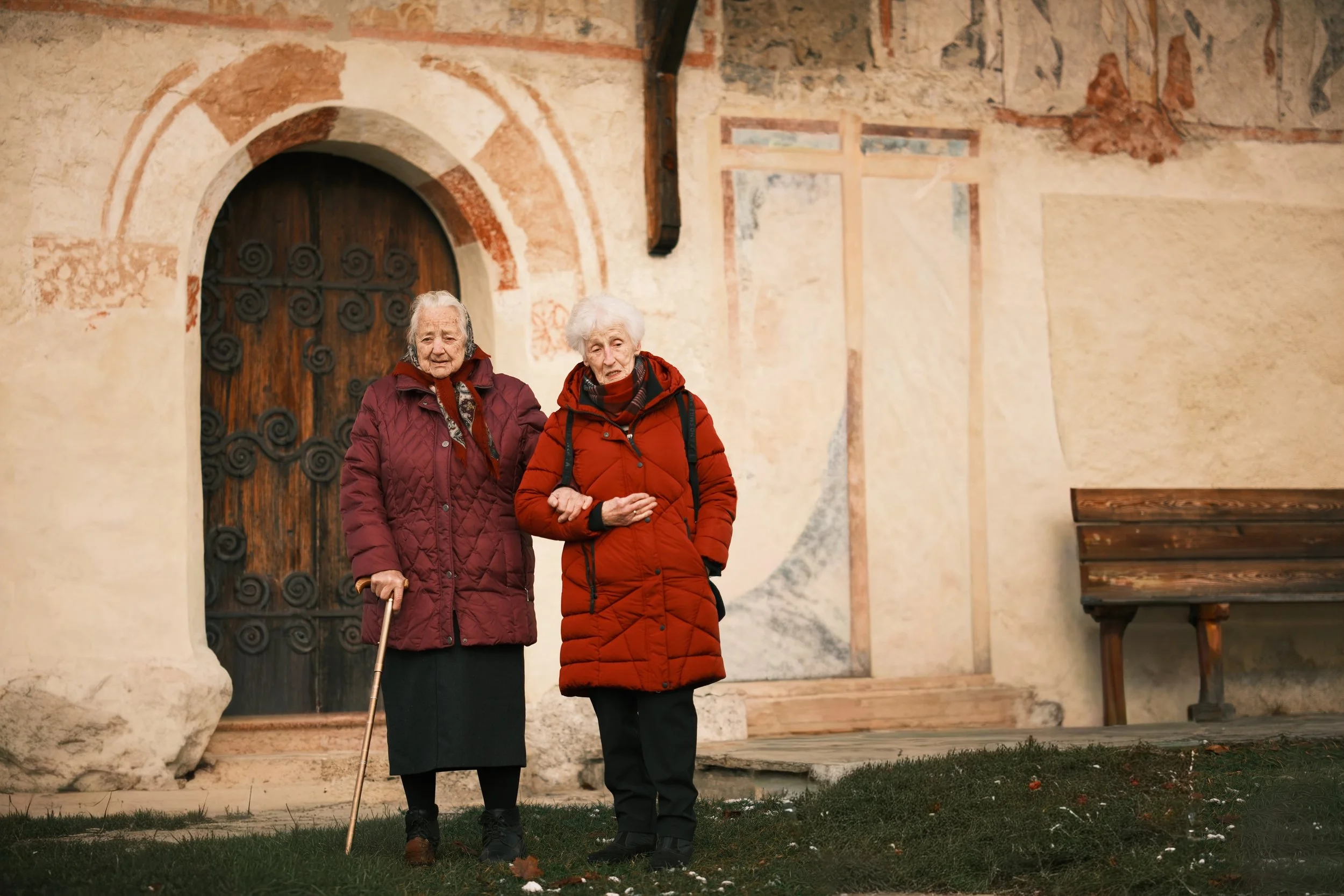 Zwei ältere Frauen in roten Jacken stehen vor einer alten, bemalten Wand, eine hält einen Gehstock, die andere arm in arm.