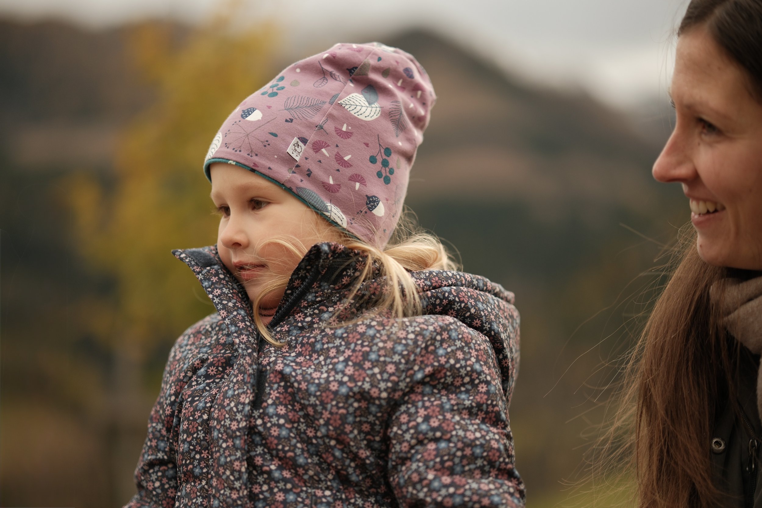 Eine junge Frau spricht mit einem kleinen Mädchen im Freien, beide tragen warme Kleidung und sind in einem herbstlichen Umfeld.