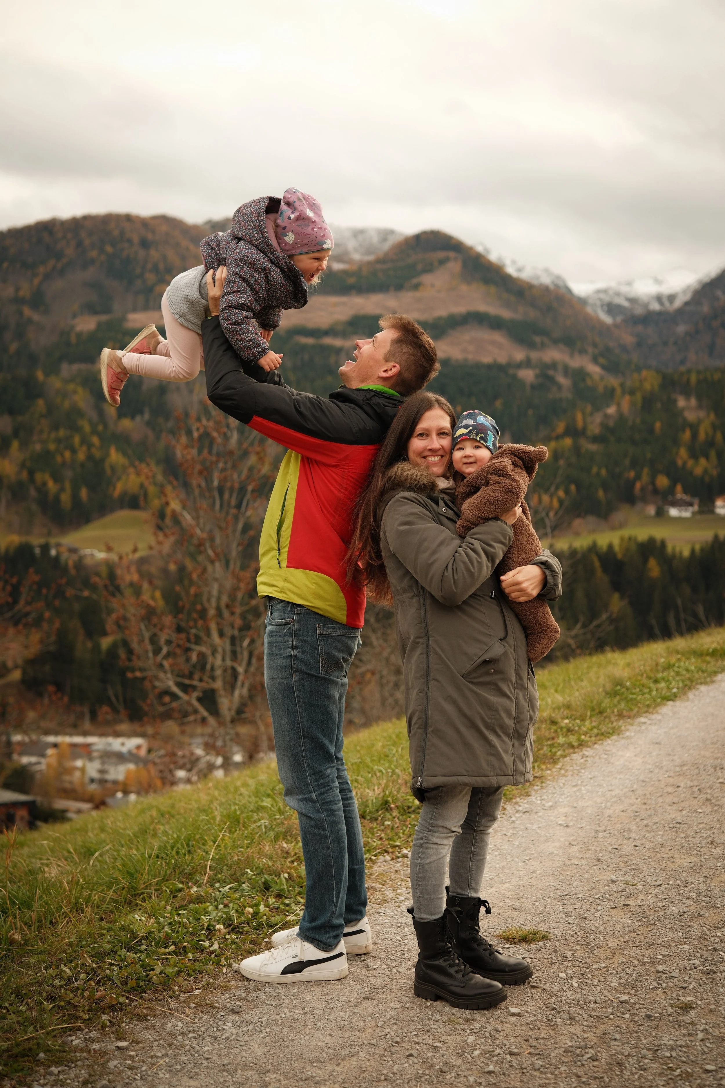 Familie mit zwei kleinen Kindern, die draußen in den Bergen bei Herbstwetter spazieren geht, der Vater hält ein Mädchen hoch, die Mutter hält ein anderes Kind in den Armen.