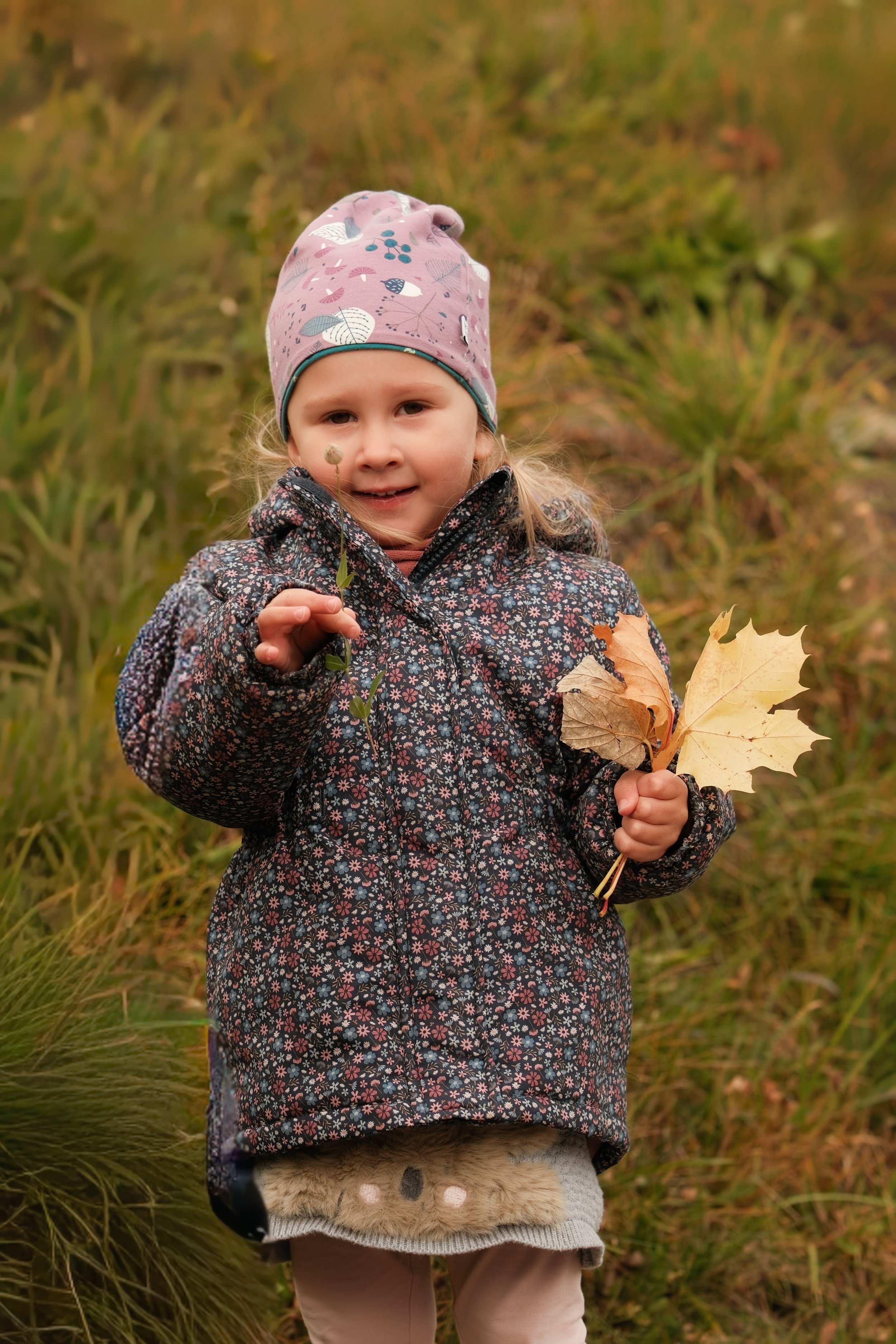Ein junges Mädchen in einem blumigen Mantel und einer lila Mütze, das im Herbst auf einer Wiese mit gelben und grünen Pflanzen steht, hält Blätter und eine kleine Pflanze in den Händen.
