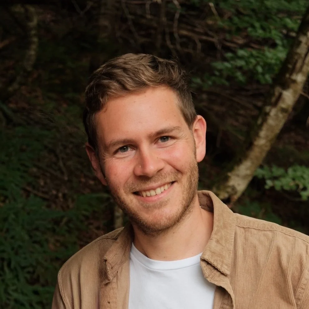 Junger Mann mit braunem kurzhaarigem Haar und weißem T-Shirt, lächelnd in einem Wald bei Tageslicht.