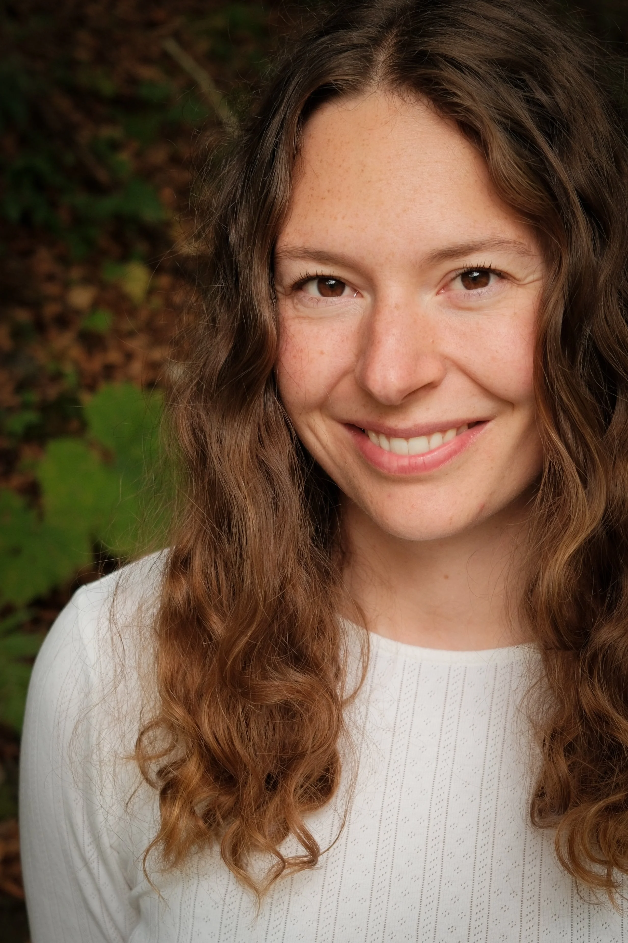 Nahaufnahme eines lachenden jungen Mädchens mit lockigem braunem Haar vor einem grünen Blätter Hintergrund.