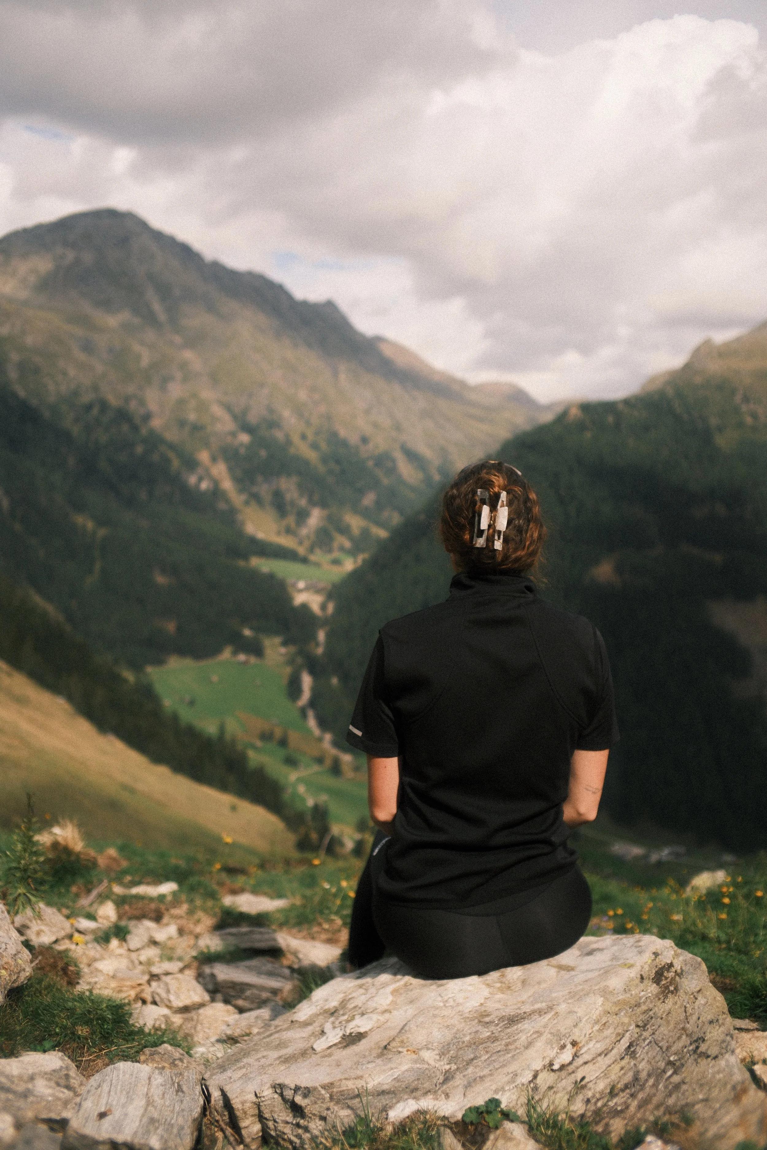 Eine Person sitzt auf einem großen Felsen und blickt auf eine grüne, bergige Landschaft mit Flüssen und bewaldeten Hügeln.