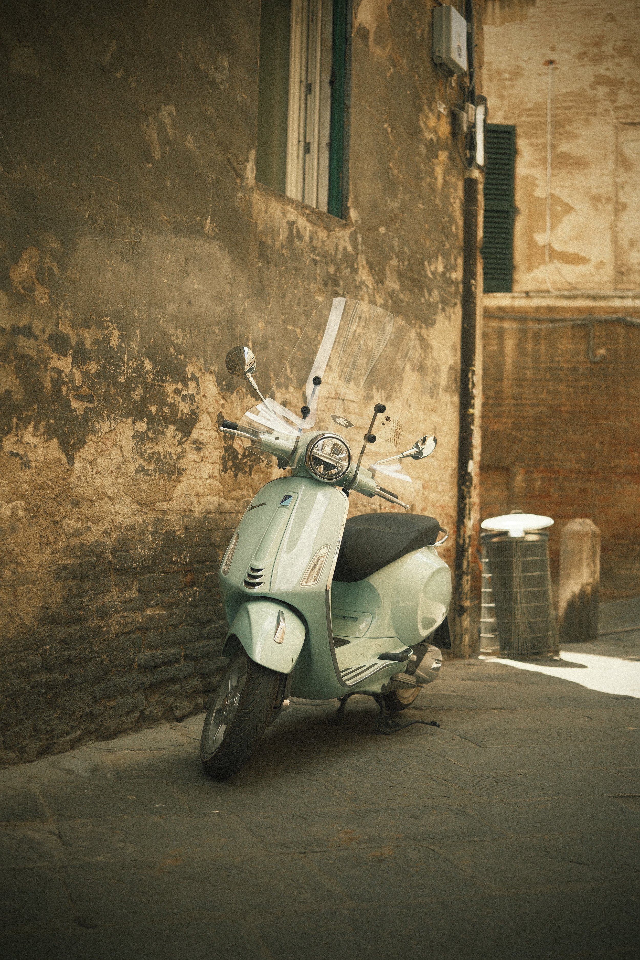 Ein vintage grüner Scooter steht an einer alten, verwitterten Steinmauer in einer engen Gasse, im Hintergrund ein Müllkorb.