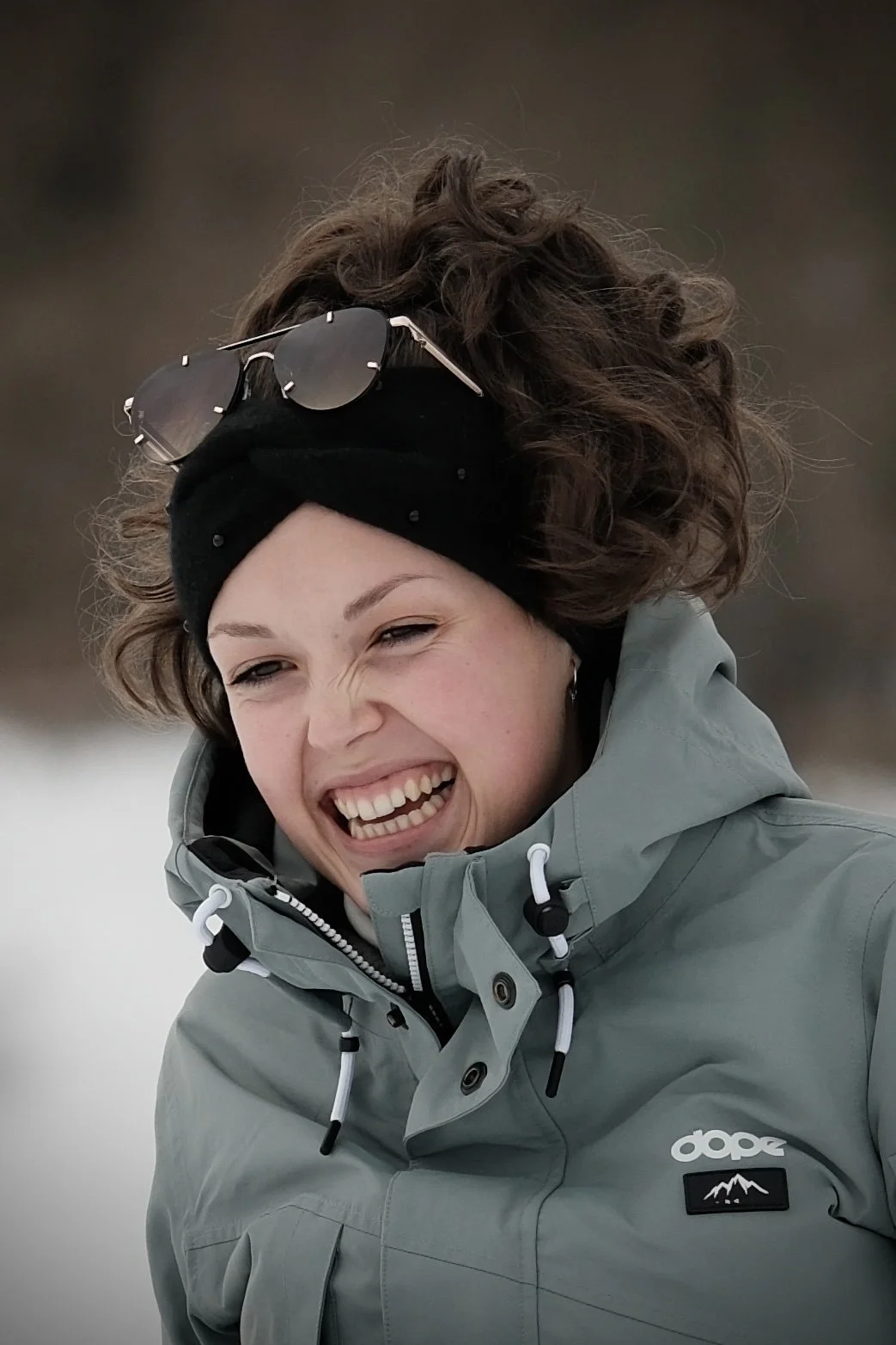 Eine lachende Frau trägt eine graue Jacke, eine schwarze Haube und hat eine Sonnenbrille auf dem Kopf.