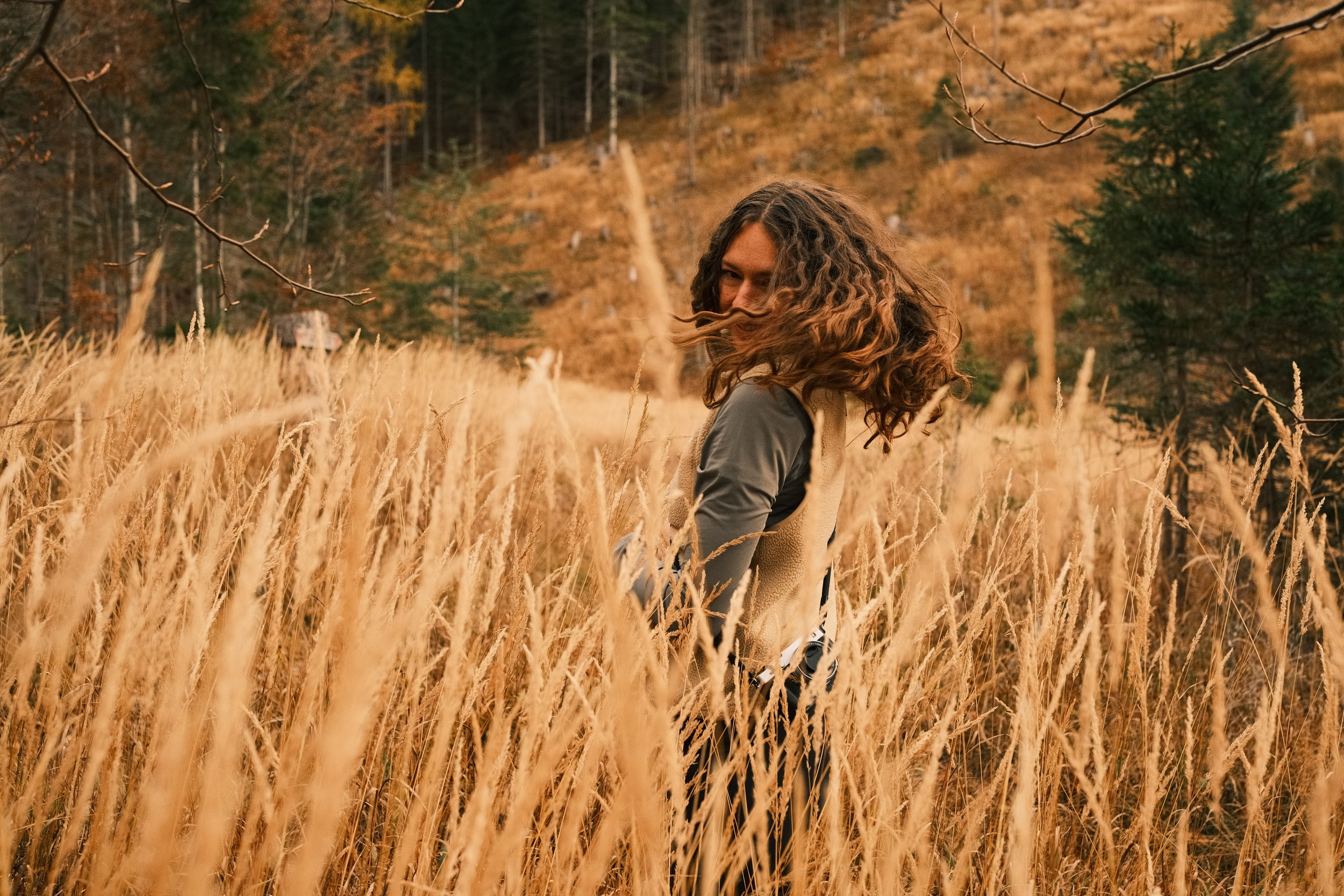 Eine Frau mit lockigen braunen Haaren steht in einem hohen, goldenen Grasfeld in einer Herbstlandschaft, umgeben von Bäumen und Bergen.