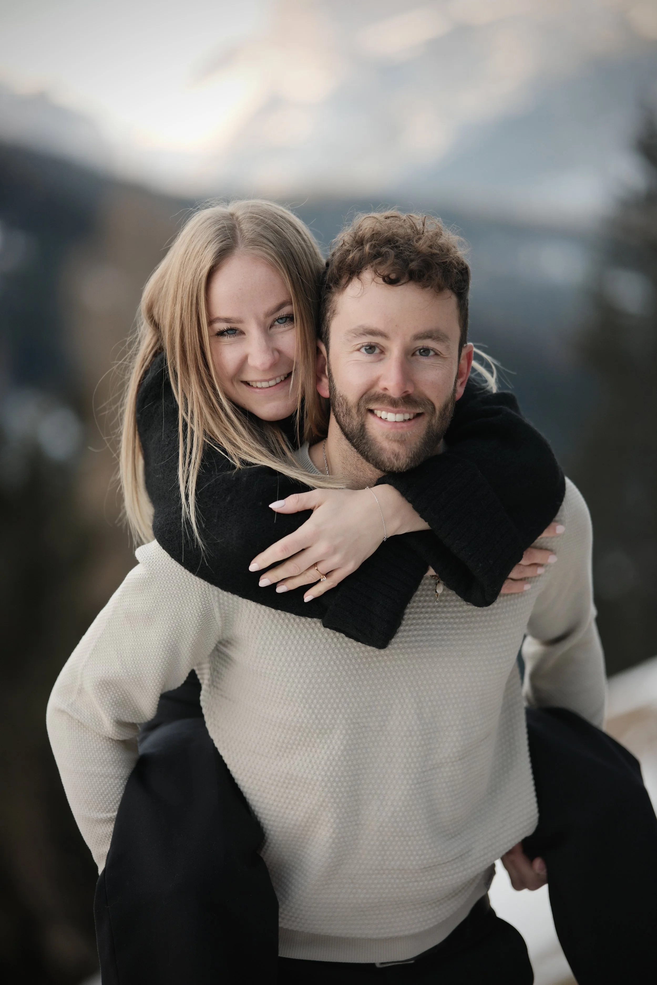 Ein lachendes Paar im Freien, der Mann trägt einen hellen Pullover, die Frau umarmt ihn von hinten, im Hintergrund Berge und Himmel.
