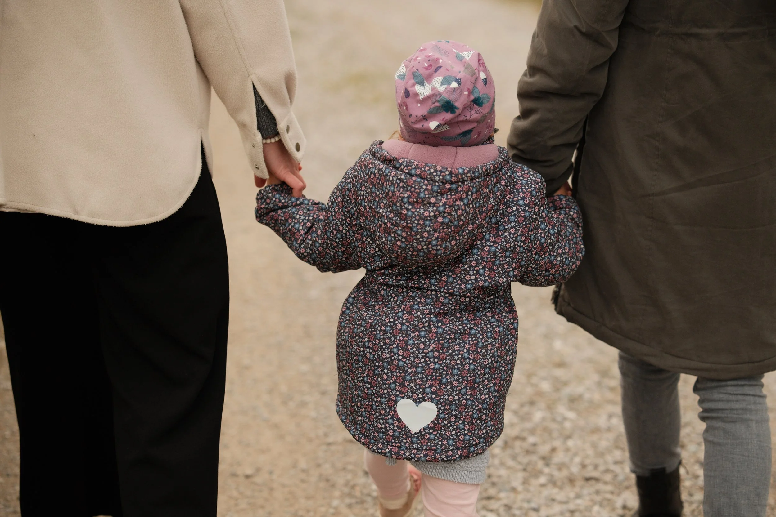 Ein kleines Mädchen wird an den Händen von zwei Erwachsenen gehalten, während sie gemeinsam einen Weg entlanggehen. Das Mädchen trägt eine dicke Jacke mit Blumenmuster und einen pinkfarbenen Hut.