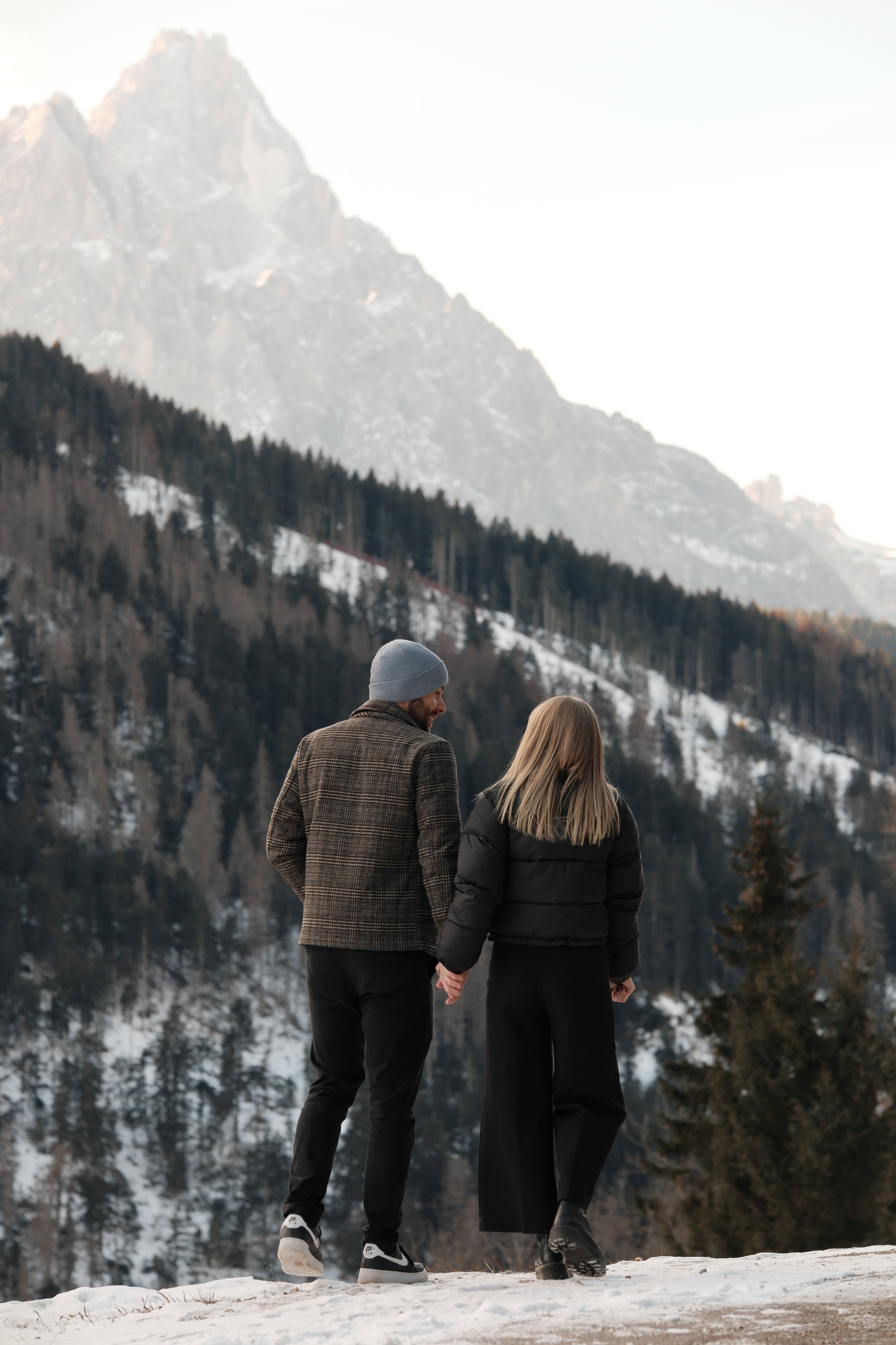 Ein Mann und eine Frau halten Händchen, während sie in winterlicher Berglandschaft spazieren gehen.