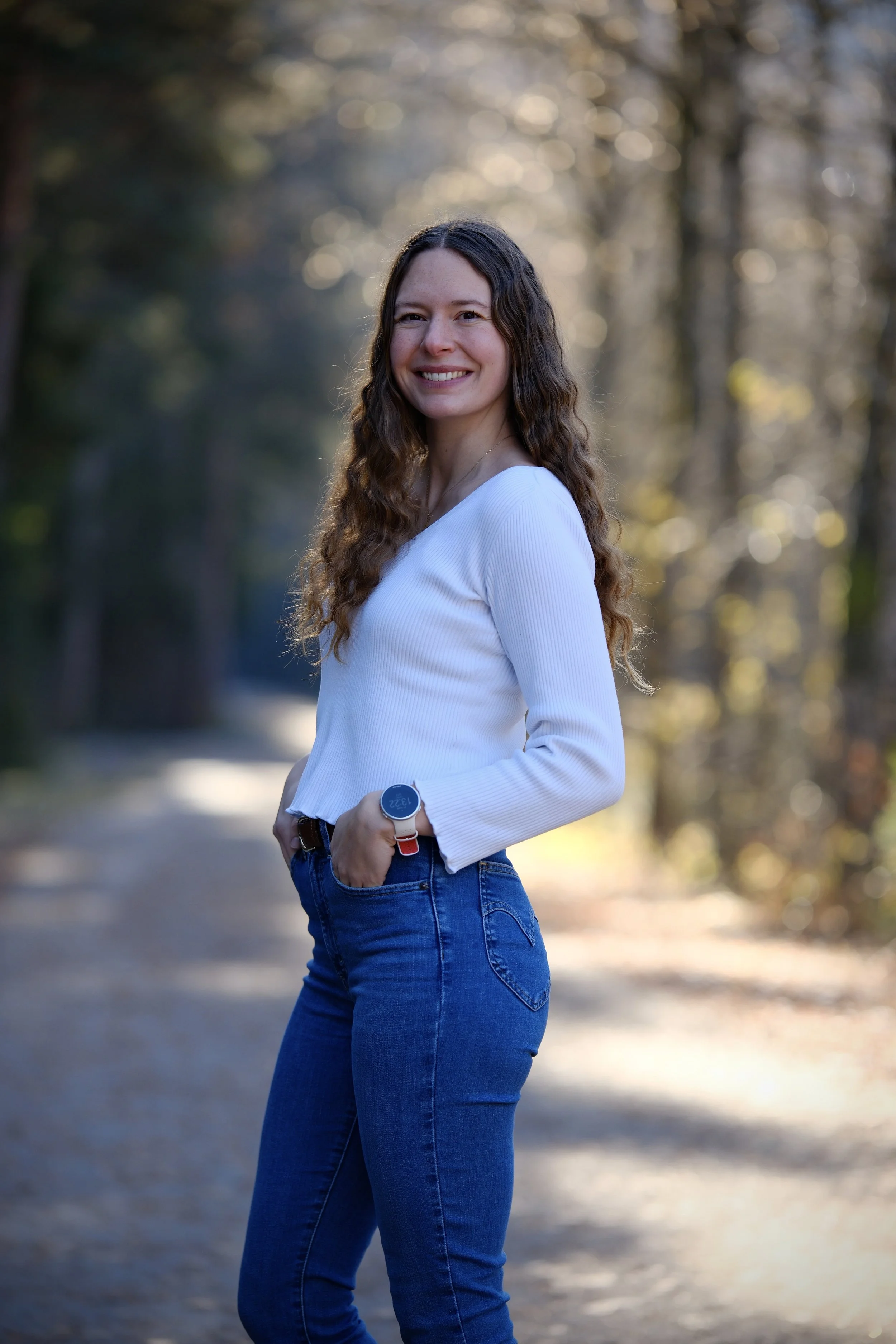 Junge Frau mit braunen, gewellten Haaren in weißem Pullover und blauen Jeans, steht auf einem Waldweg bei Sonnenuntergang.