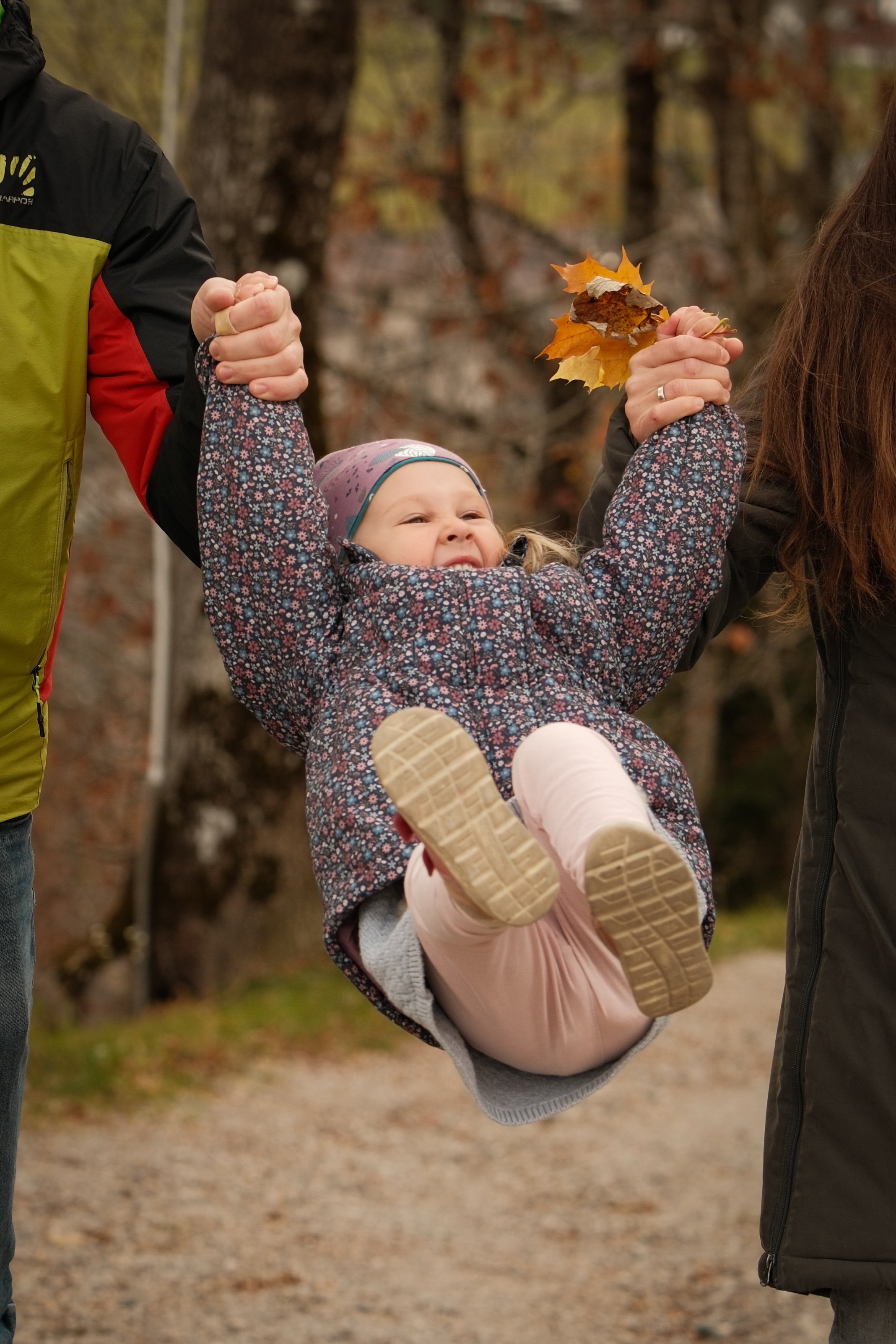 Ein kleines Mädchen wird von zwei Erwachsenen in einem Park gehalten, sie schwingt fröhlich in der Luft und hält herbstliches Laub in der Hand.