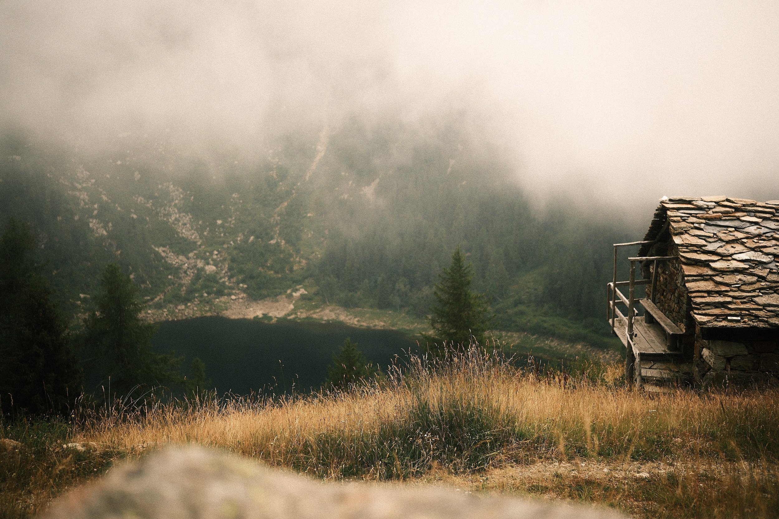 Ein steinernes Haus auf einem Hügel mit einer hölzernen Terrasse, umgeben von hohem Gras und Bäumen, mit einem See im Hintergrund und Bergen, die teilweise von Nebel bedeckt sind.