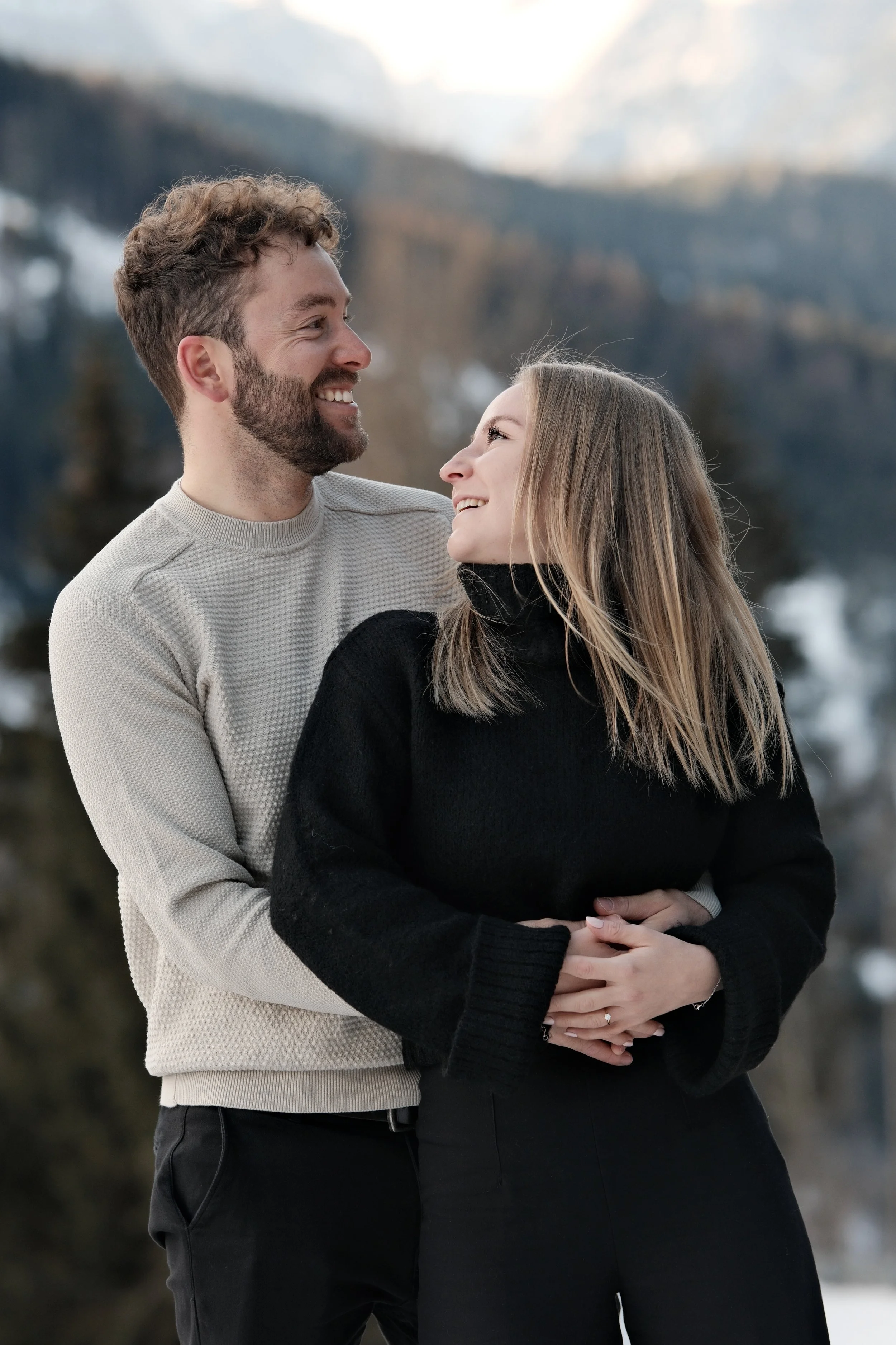 Ein lachendes Paar umarmt sich in einer Winterlandschaft, mit Bergen im Hintergrund.