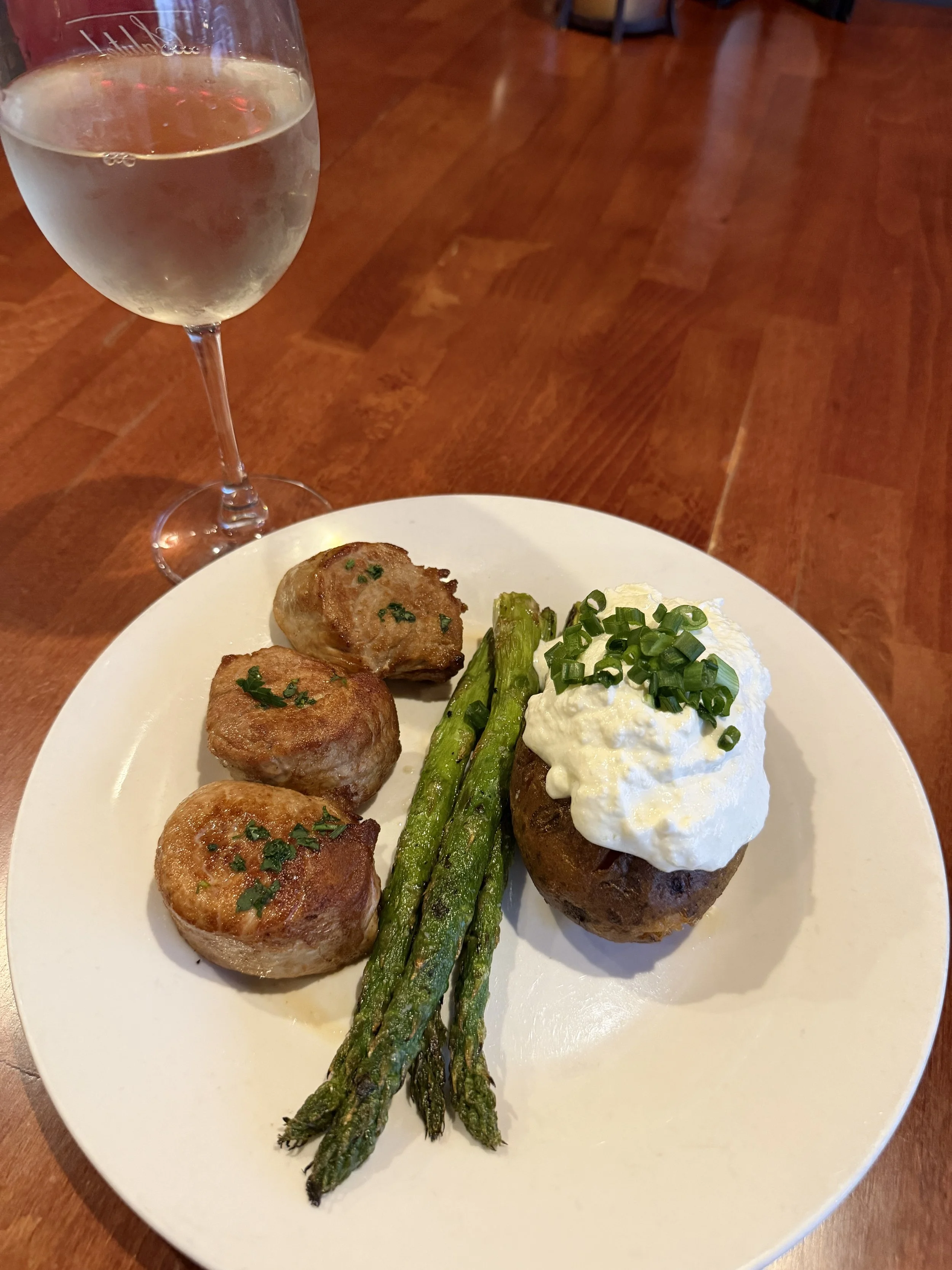 A white plate with three roasted potatoes topped with chopped green herbs, three steamed asparagus spears, and a baked potato with sour cream and chopped green onions. A glass of white wine is placed nearby on a wooden table.