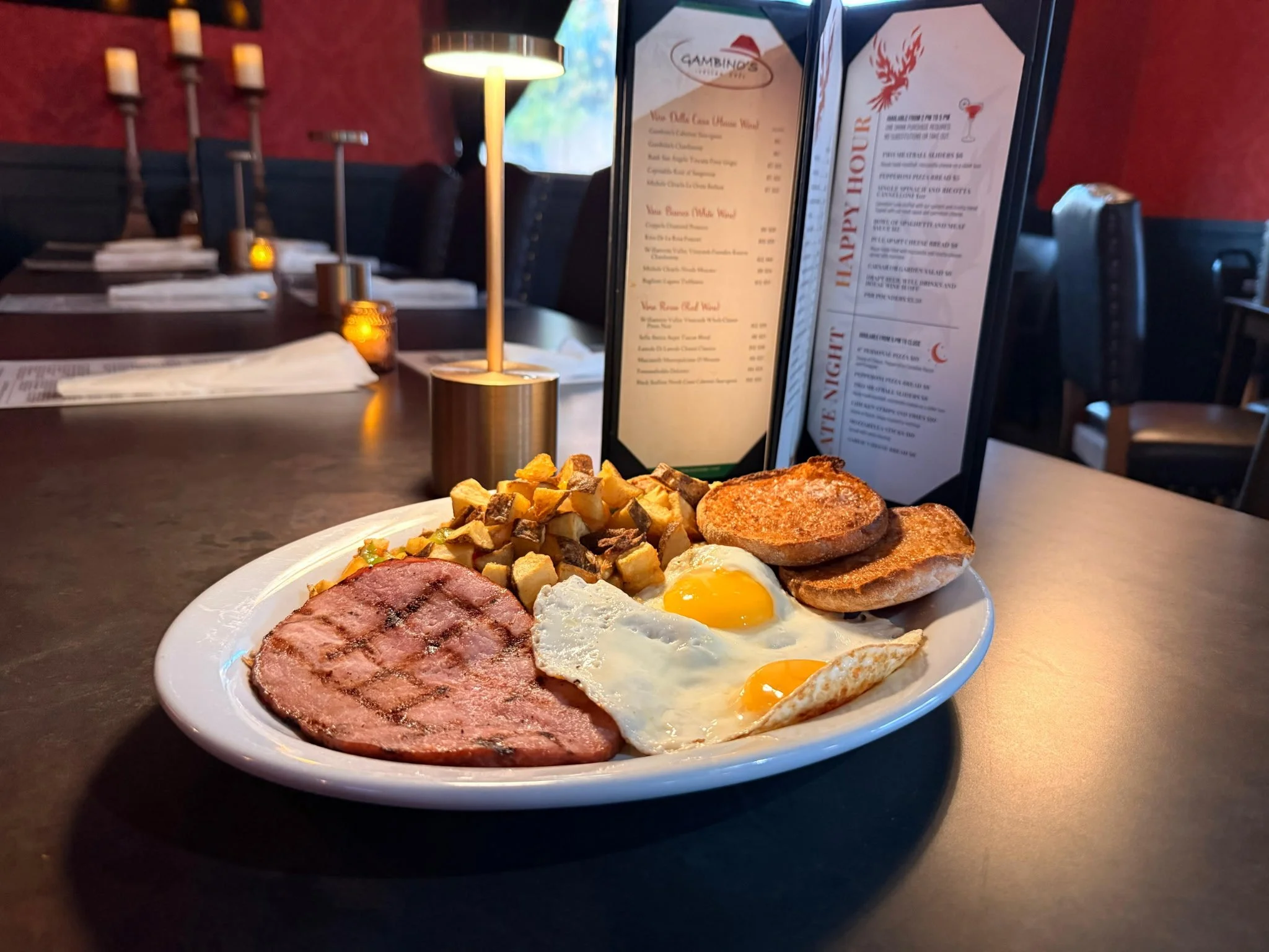 A breakfast plate with two sunny-side-up eggs, grilled ham, roasted potatoes, and toasted bread slices on a dark restaurant table.