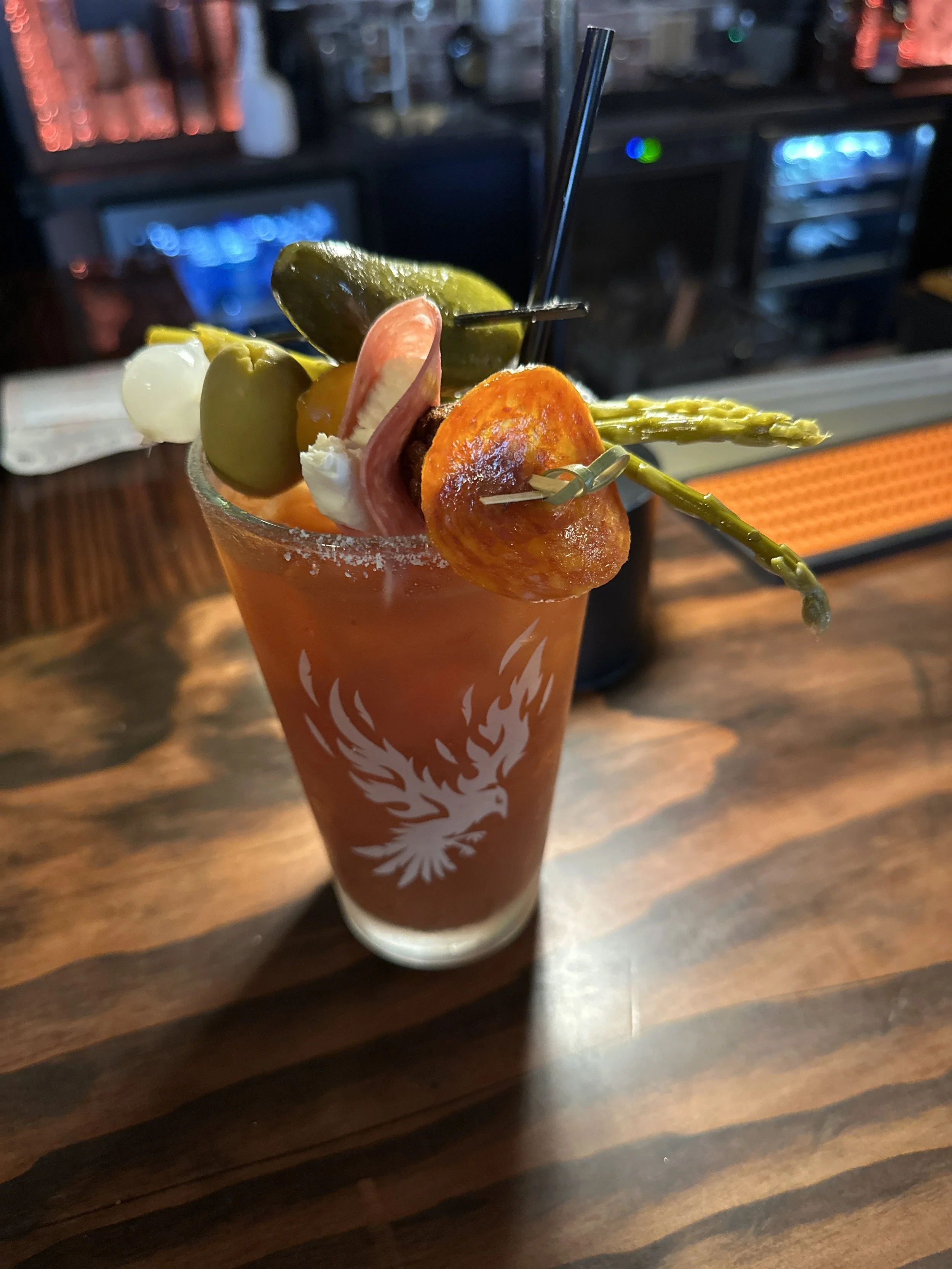 A Bloody Mary cocktail garnished with a pickle, green olives, a slice of pepperoni, a cherry, an asparagus spear, and a lemon wedge, sitting on a wooden bar counter.