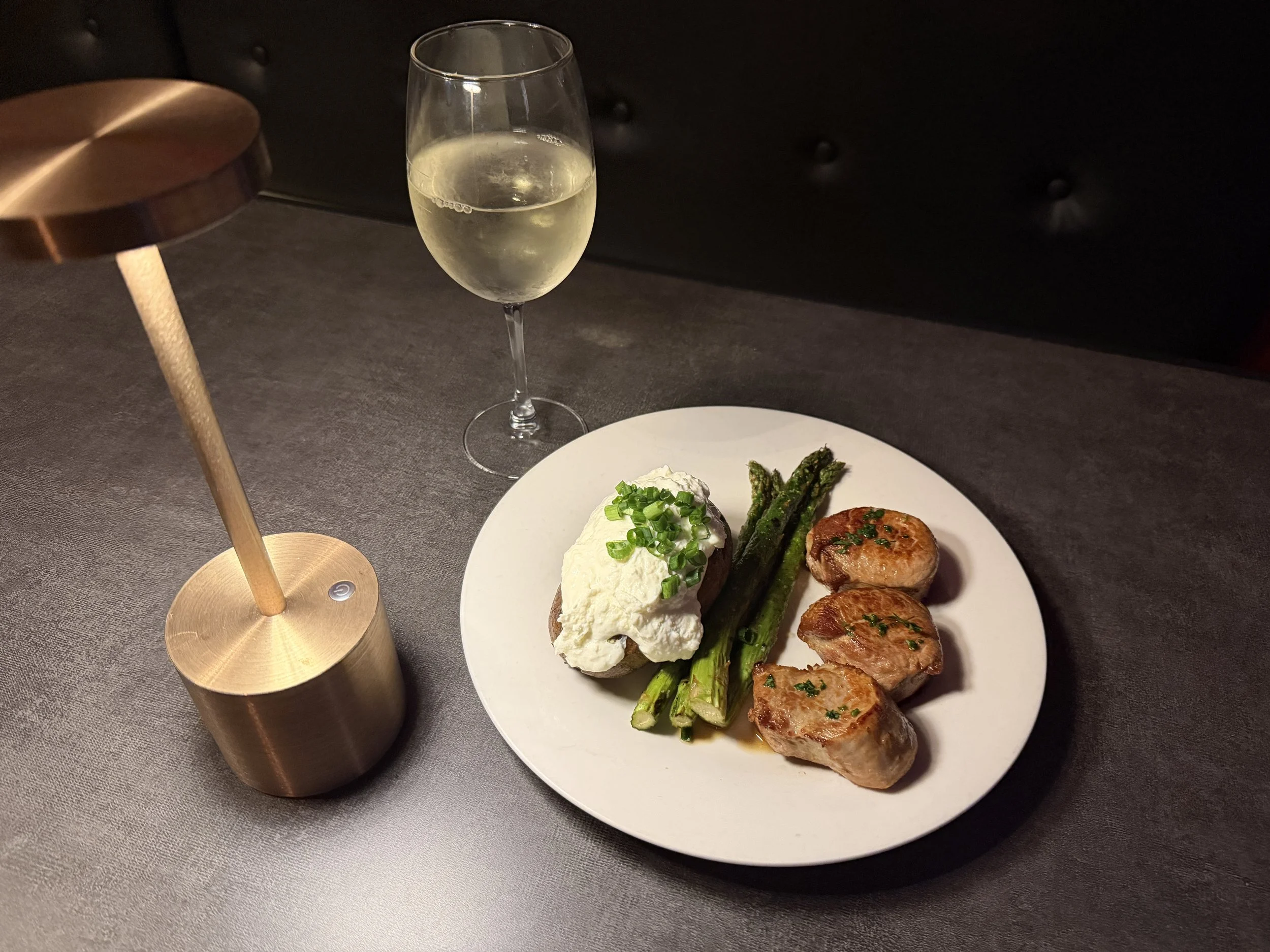 A white plate with three cooked scallops topped with herbs, grilled asparagus spears, and a baked potato topped with whipped cream and chopped green onions. A glass of white wine is in the background, on a dark table with a copper-colored table lamp.