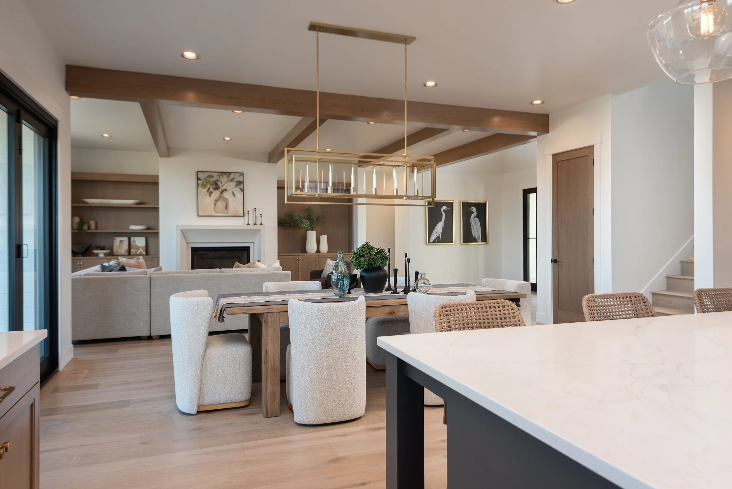 Open-concept living and dining area with a modern farmhouse design, featuring a dining table with cream upholstered chairs, a neutral-toned living room with a fireplace, wooden beams on the ceiling, decorative wall art, and contemporary lighting fixtures.