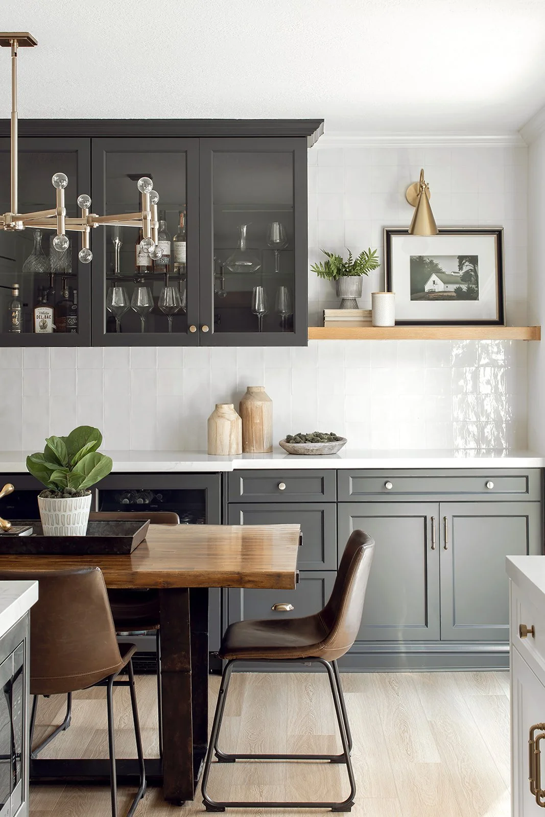 Modern kitchen with dark gray cabinets, glass-front upper cabinets with glassware, a wooden dining table, brown leather chairs, decorative vases, a framed photo, a small potted plant, and a gold wall-mounted light fixture.