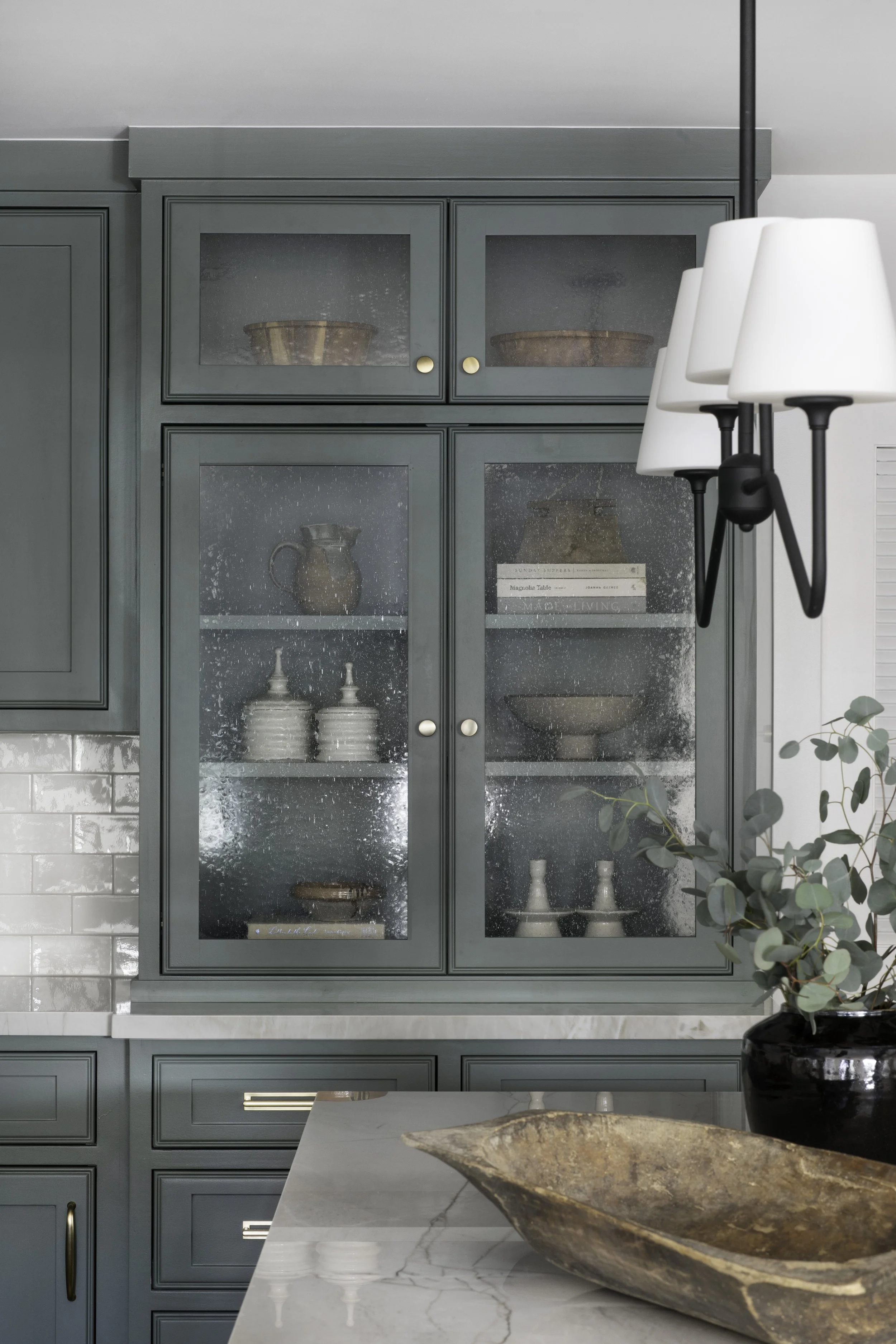 A kitchen with green cabinets behind a marble countertop, a black and white wall-mounted light fixture, decorative pottery and books inside a glass-front cabinet, and a large wooden bowl on the countertop.