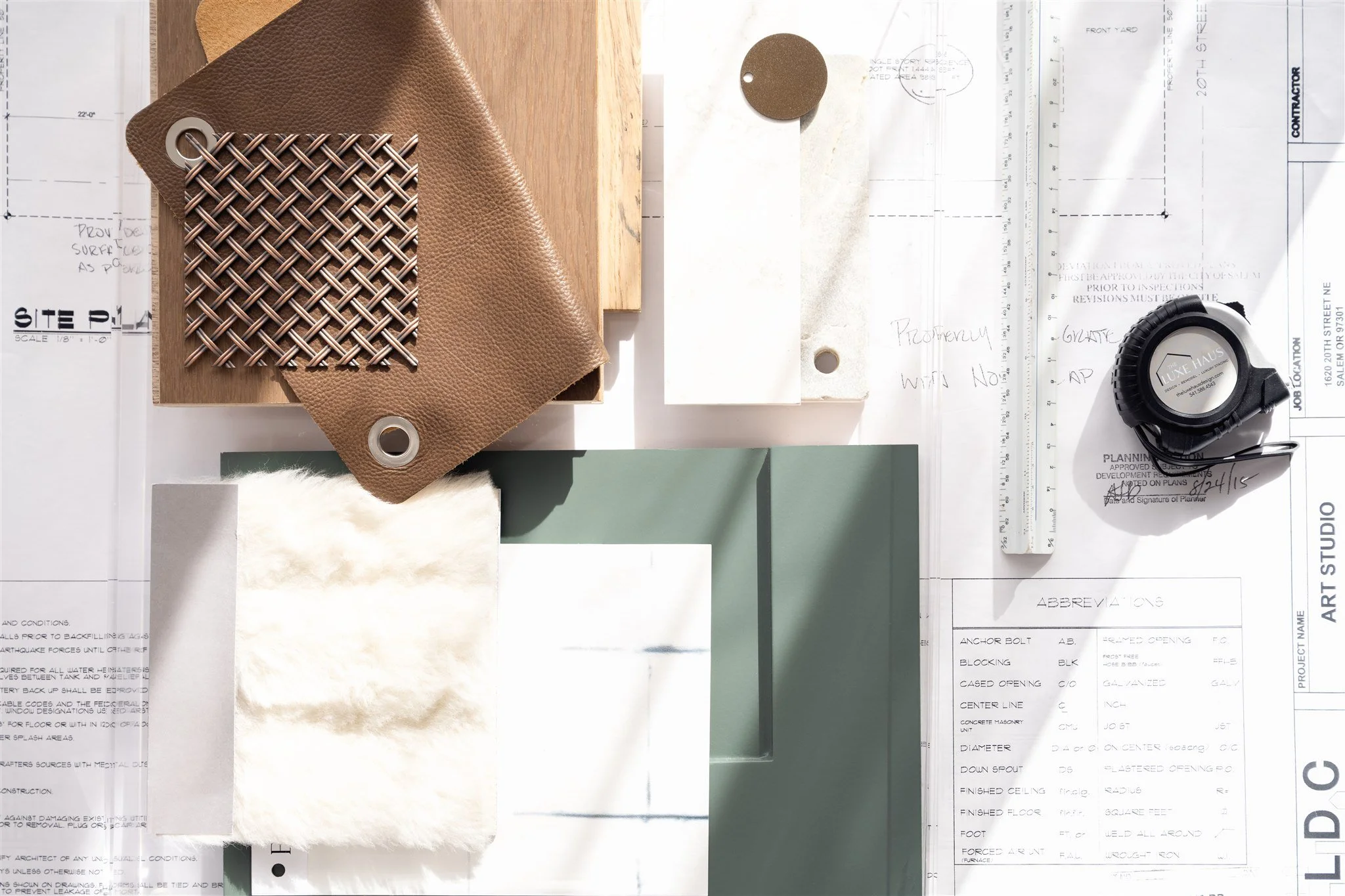 A collection of interior design samples and tools on architectural plans, including fabric swatches, paint chips, a measuring tape, and drafting tools.