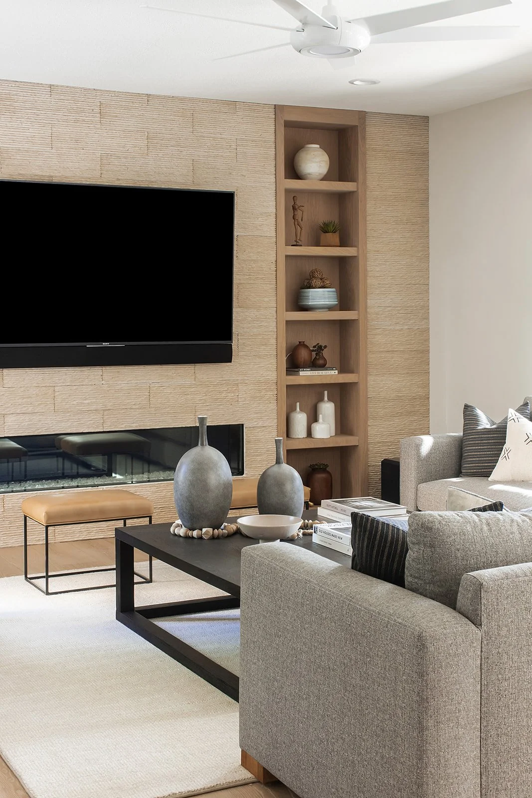 Modern living room with beige textured wall, mounted flat-screen TV, built-in fireplace, wooden shelving with decorative vases, and a gray sofa with black and white pillows. Coffee table with vases and books, cream rug, ceiling fan.