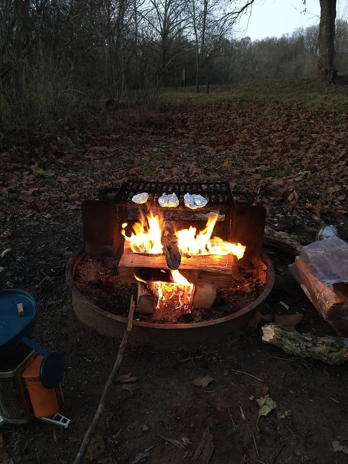 a roaring campfire with foil wrapped food on a grate. a bundle of firewood is to the side, and fallen leaves extend away in the distance