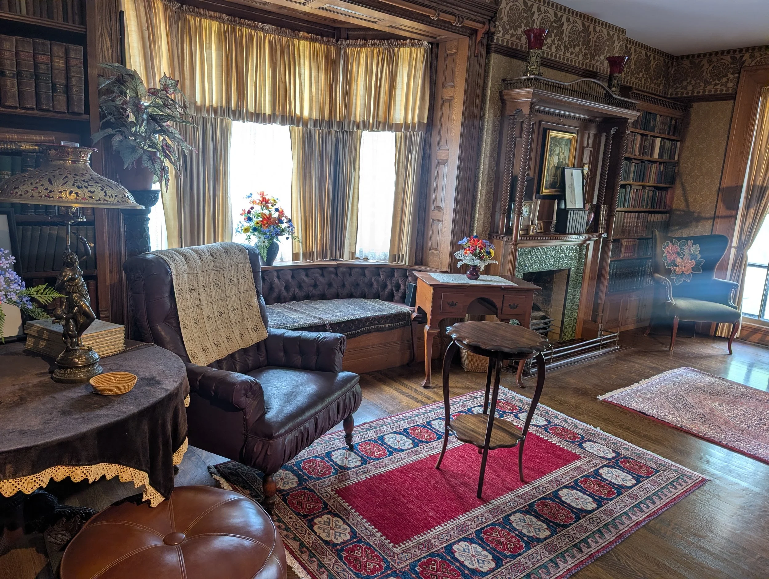 a victorian era library with a window seating area and two other chairs prominent. a small circular table is in the center of the room. the hardwood floors have rectangular "oriential" rugs on them. a fireplace has books on the mantle.