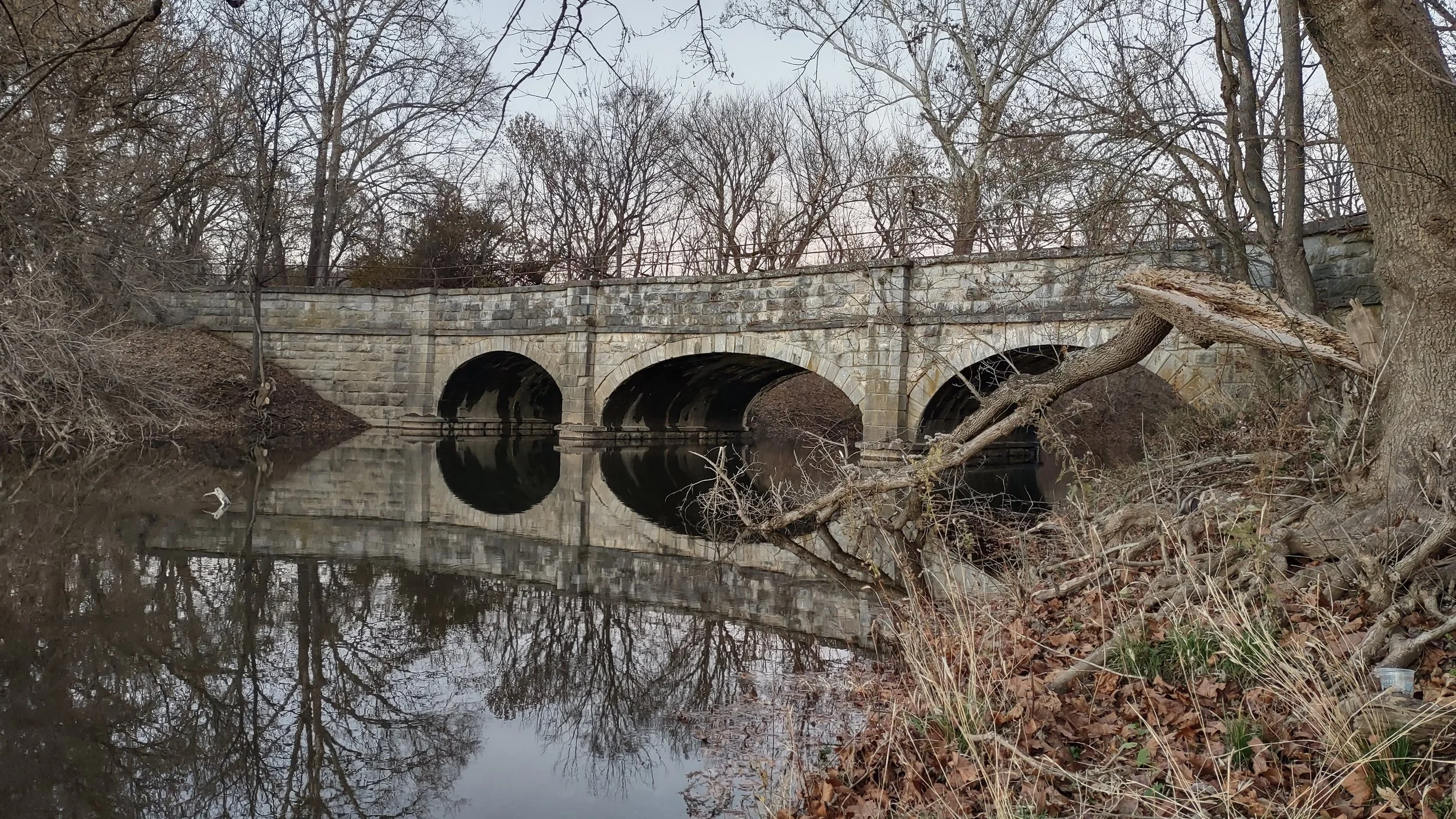 The Antietam Aqueduct of the C&O Canal Towpath, over Antietam Creek. The view is from the downstream side of the canal; the photographer is standing almost at the confluence of the creek and the Potomac River. The trees are mostly leafless.