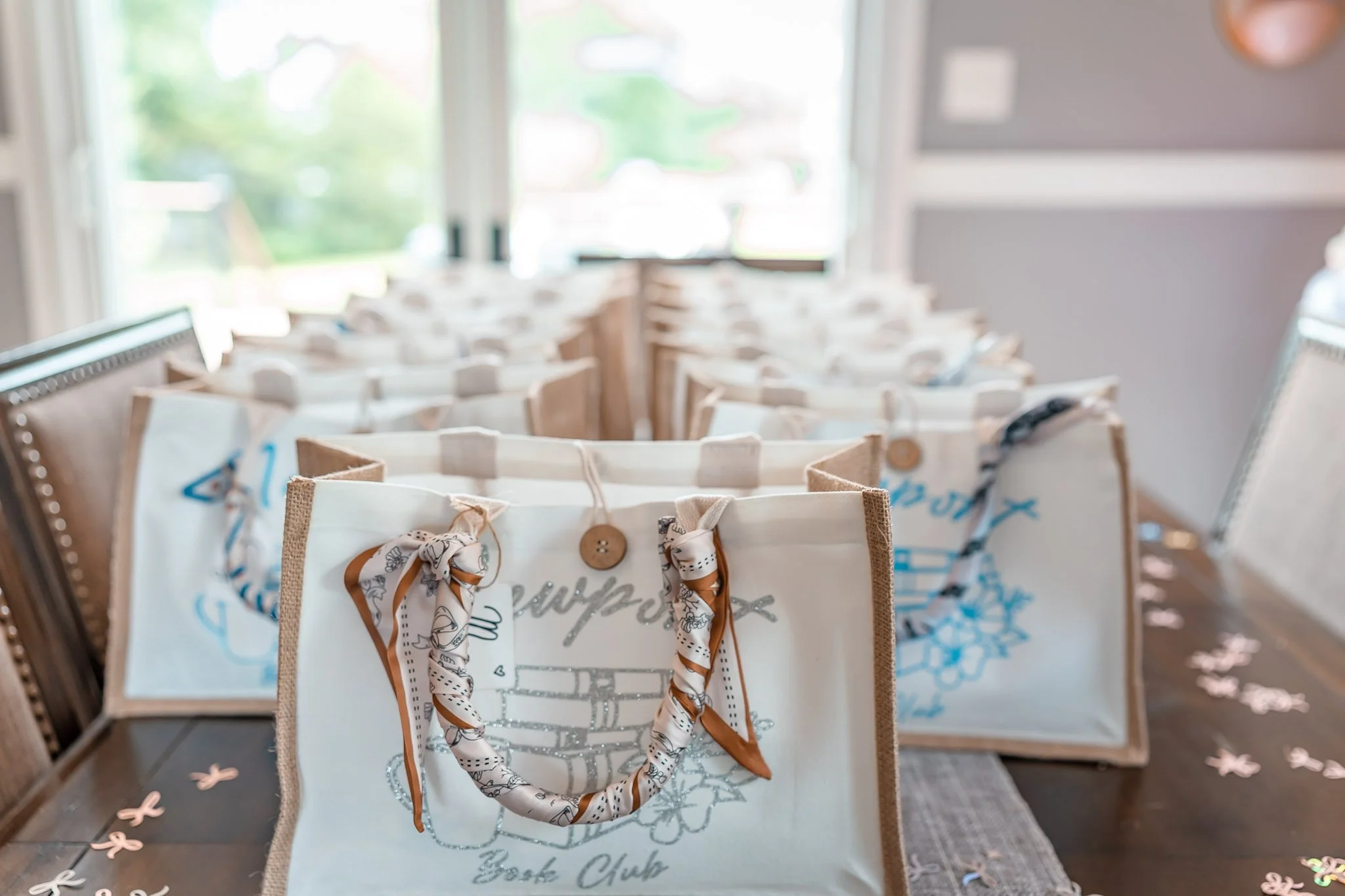 Decorative tote bags with embellished handles and embroidered designs, arranged on a dining table near a window, with confetti scattered on the surface.